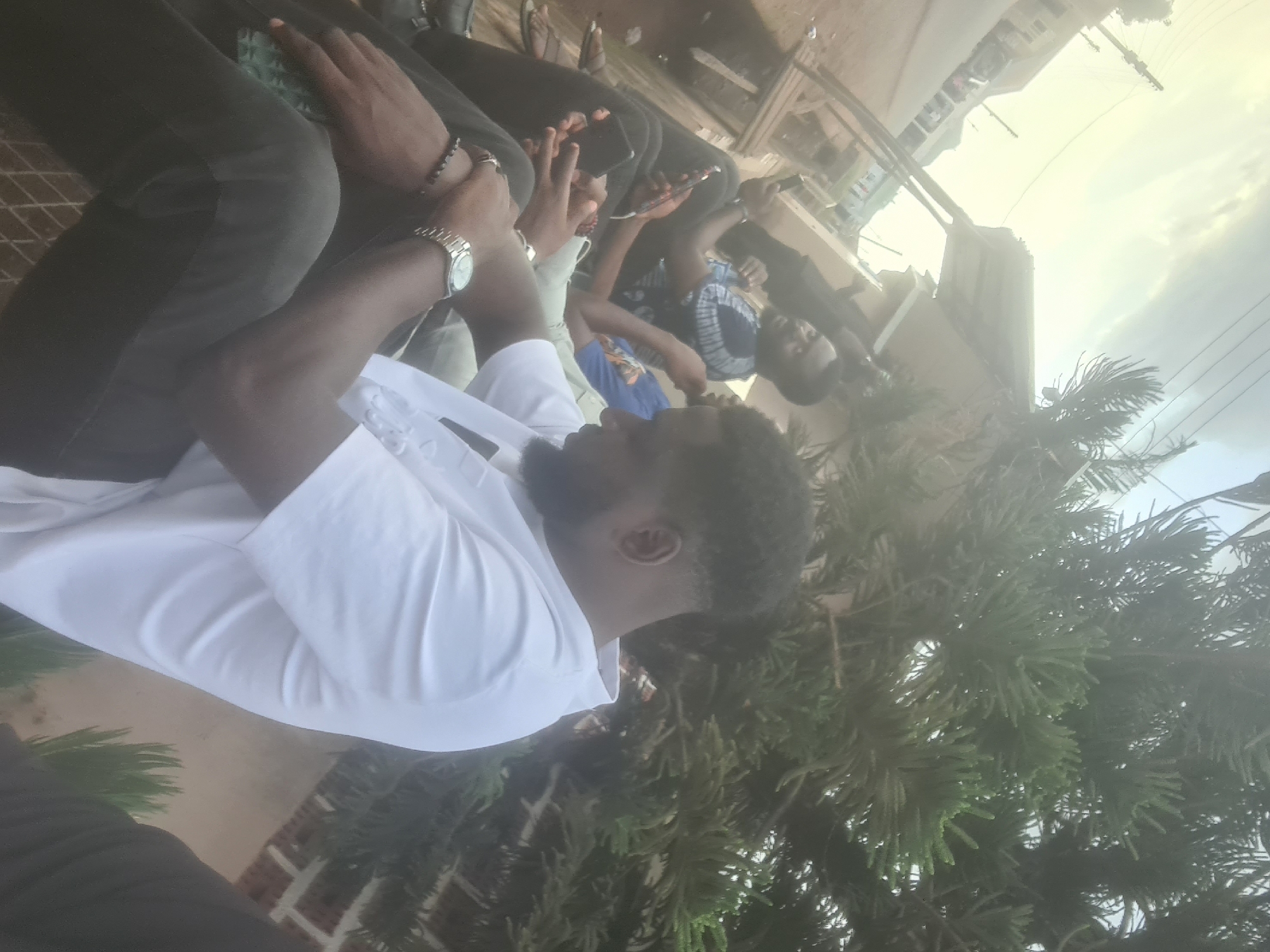 A group of people are seated together outside, possibly in Jos, Nigeria. One individual, closest to the viewer, is wearing a white shirt and a watch on their left wrist. Others in the group are using mobile phones. The background includes buildings, utility lines, and trees, suggesting an urban environment. The image appears to have been taken at an angle.