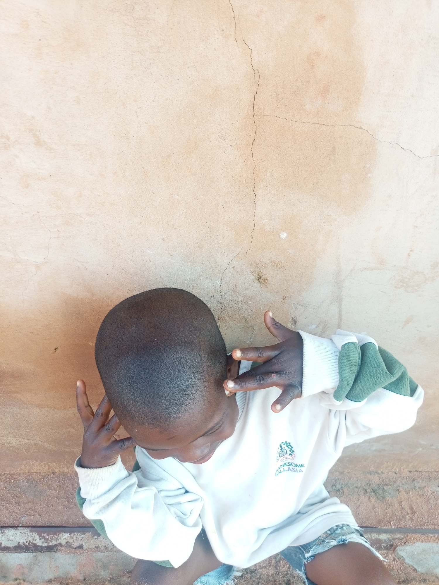 An overhead shot shows a young child looking down with their right hand resting on their ear and their left hand in front of their face in the city of Tempure, Nigeria. The child is wearing a light-colored sweatshirt with green accents on the sleeves and ripped denim shorts. They are positioned against a textured, light-brown wall with visible cracks. The ground beneath the child appears to be made of gravel and dirt.