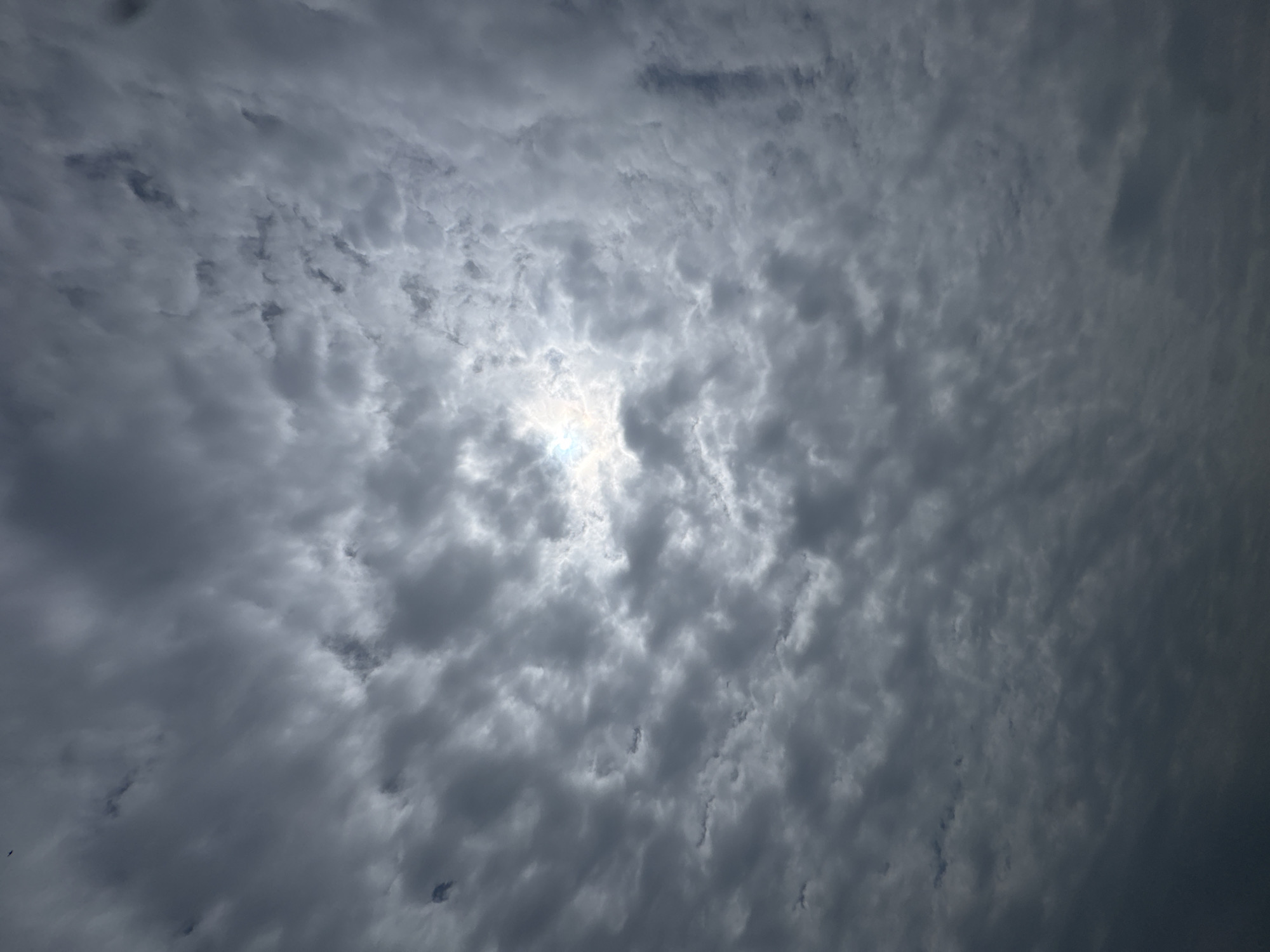 The image shows a sky filled with dark, overcast clouds. The sun is partially visible through a break in the clouds, creating a bright, hazy area. The clouds appear to be thick and textured, with varying shades of gray and white. The overall impression is of a cloudy, potentially stormy day. No people, subjects, objects, or actions are visible in the image. The location is unknown.
