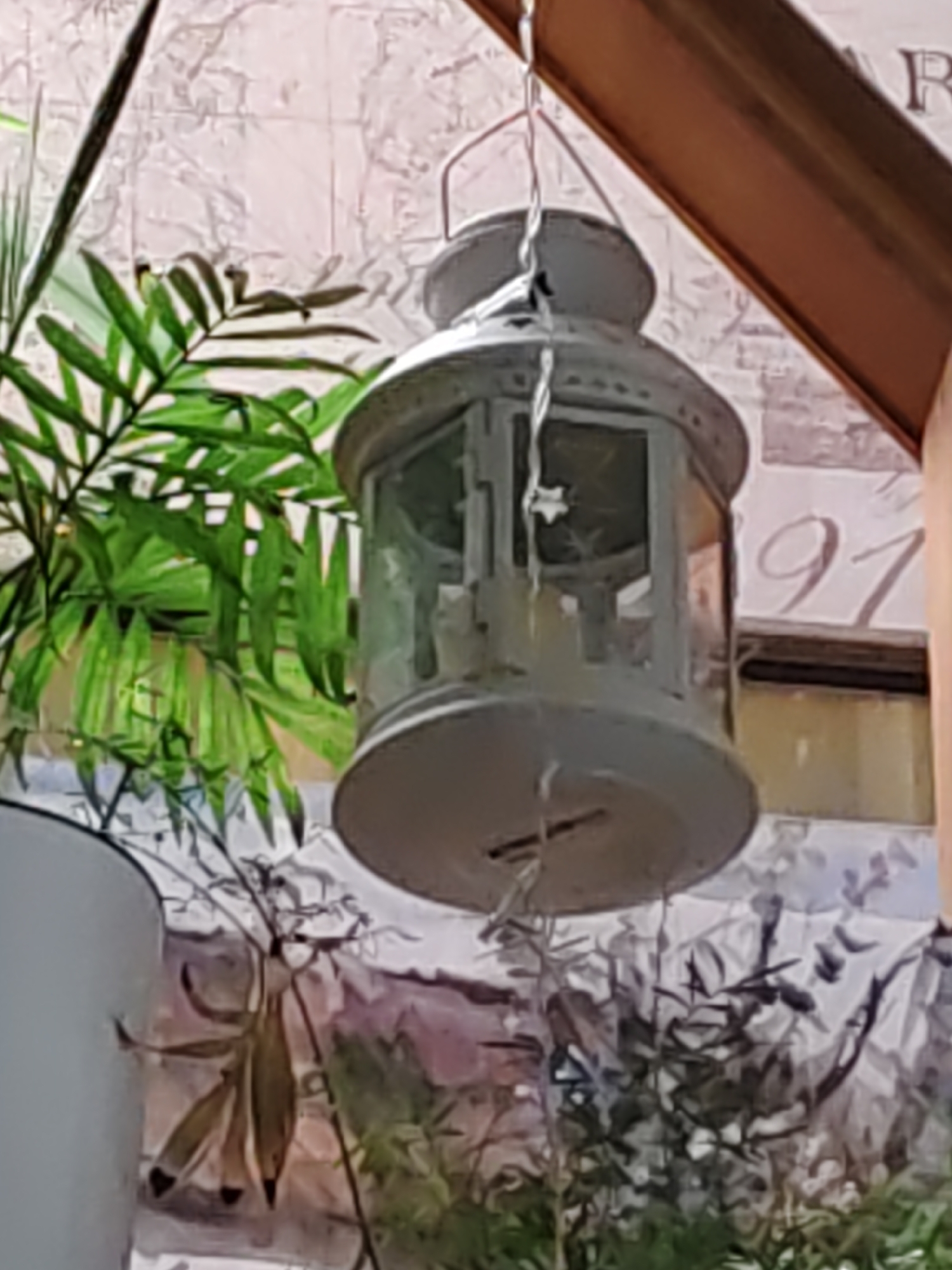 A close-up, slightly out-of-focus shot features a vintage-style, white metal lantern hanging from a wire. The lantern has glass panes and appears to have candle holders inside. It is positioned in front of a textured wall with a floral or marbled pattern. To the left of the lantern, a portion of a green plant with frond-like leaves is visible, and to the right, a wooden beam is angled downwards. In the background, the wall features decorative elements, including what appears to be the number "91" in script. The lighting suggests an indoor or sheltered outdoor setting. The image lacks specific identifiers for Rostov-on-Don.