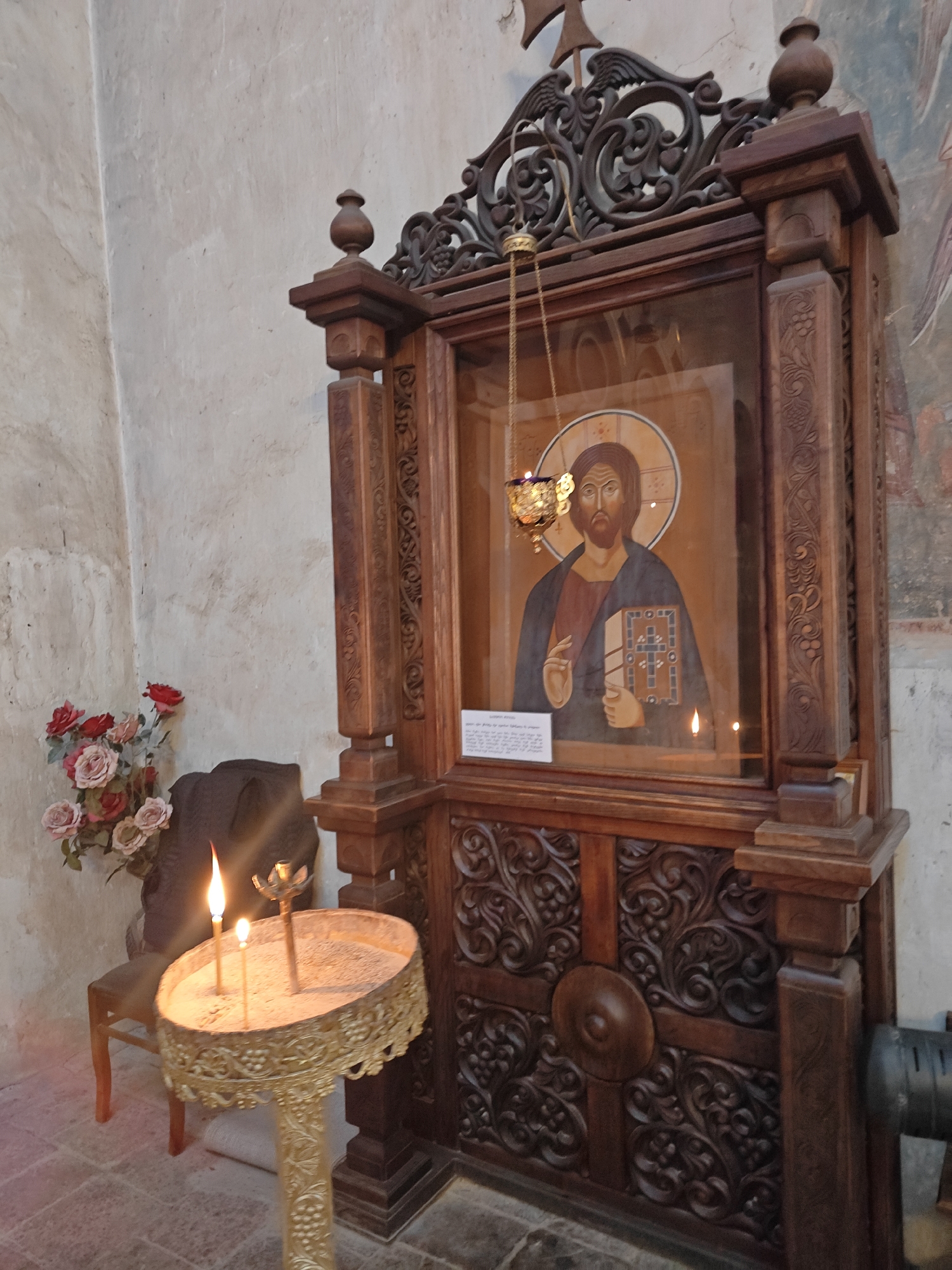 The image displays an interior scene within an Orthodox Christian church or monastery, likely in Pkhundavi, Georgia. The central feature is an elaborate, dark wooden iconostasis or shrine, richly adorned with intricate floral and scrollwork carvings.
Within the wooden structure, behind a glass panel, rests a painted icon of Jesus Christ. He is depicted with a halo, a beard, and a solemn expression, holding an open book (likely the Gospels) in his left hand while his right hand is raised in a gesture of blessing. A small, ornate golden vigil lamp (kandili) hangs directly in front of the icon, suspended by chains, its burning wick casting a warm glow.
To the left of the iconostasis, an ornate, golden-colored metal stand holds several devotional candles. Two candles are actively burning, one with a prominent flame, set in a tray filled with sand or ash. Behind this stand, a bouquet of artificial or dried roses in shades of pink and red rests on a simple dark wooden chair or stool.
The surrounding walls are light-colored and textured, suggesting stone or aged plaster, with faint traces of a painted mural visible on the right. A small, white rectangular plaque with text in an indecipherable script, presumably Georgian, is visible on the lower left portion of the icon, behind the glass. The presence of burning candles and the vigil lamp suggests an ongoing act of prayer or devotion within a sacred space, creating a serene and reverent atmosphere.