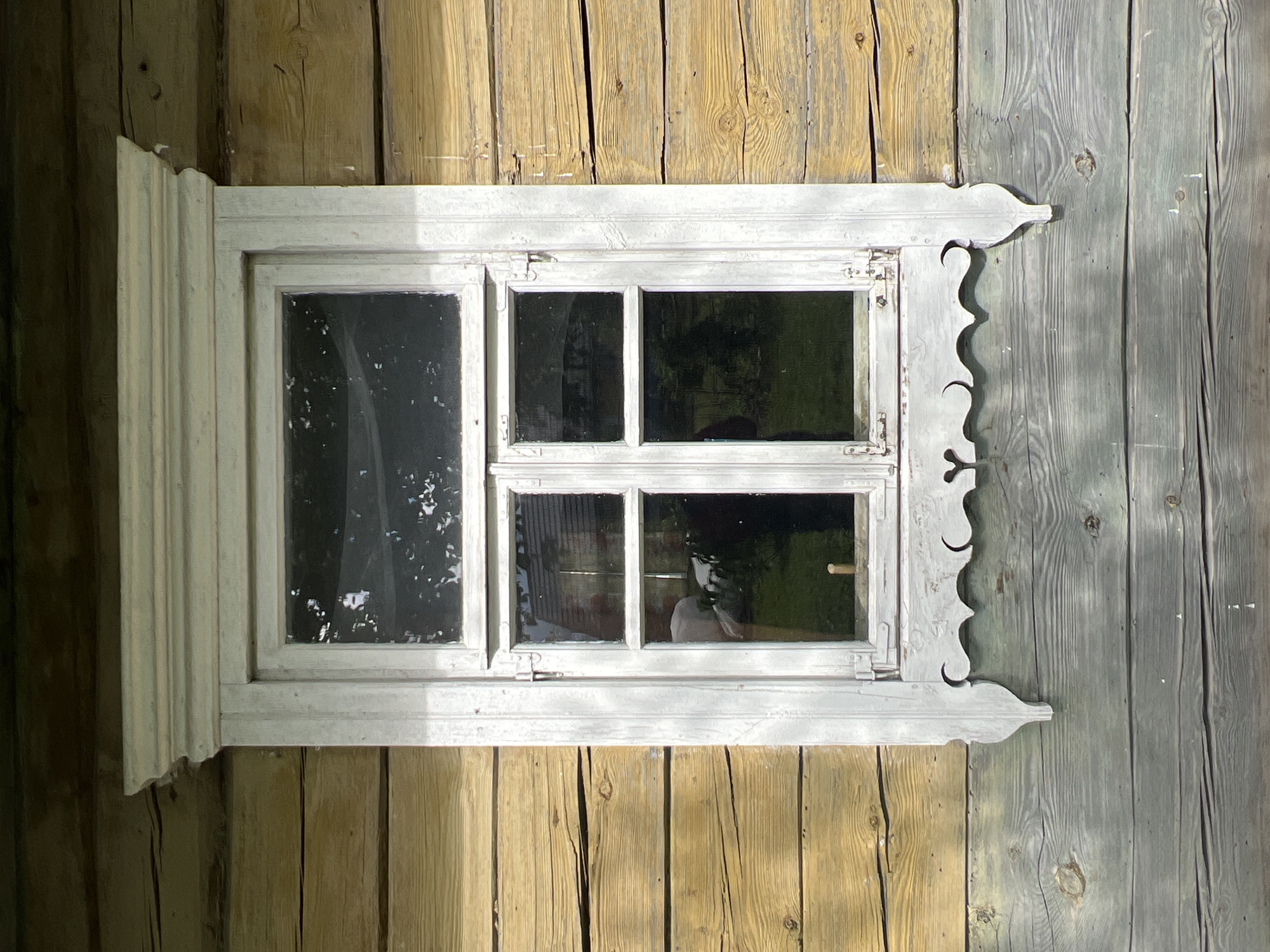 A weathered, white-painted wooden window is centered on a log wall of a traditional rural Belarusian house. The upper portion of the wall is constructed of horizontally laid, yellow-tinted logs with visible grain and occasional cracks. The lower portion of the wall is made of wider, grey-toned wooden planks, also showing natural wear and grain patterns. The window itself is divided into four panes, two on top and two on the bottom, framed by white-painted wood. The window frame features decorative elements, including a carved, scalloped trim piece below the lower panes and ornate finials on the sides. Reflections in the glass suggest an outdoor environment with trees and possibly a building visible. The scene appears to be in Бучацінскі сельскі Савет, Belarus, indicative of traditional rural architecture.