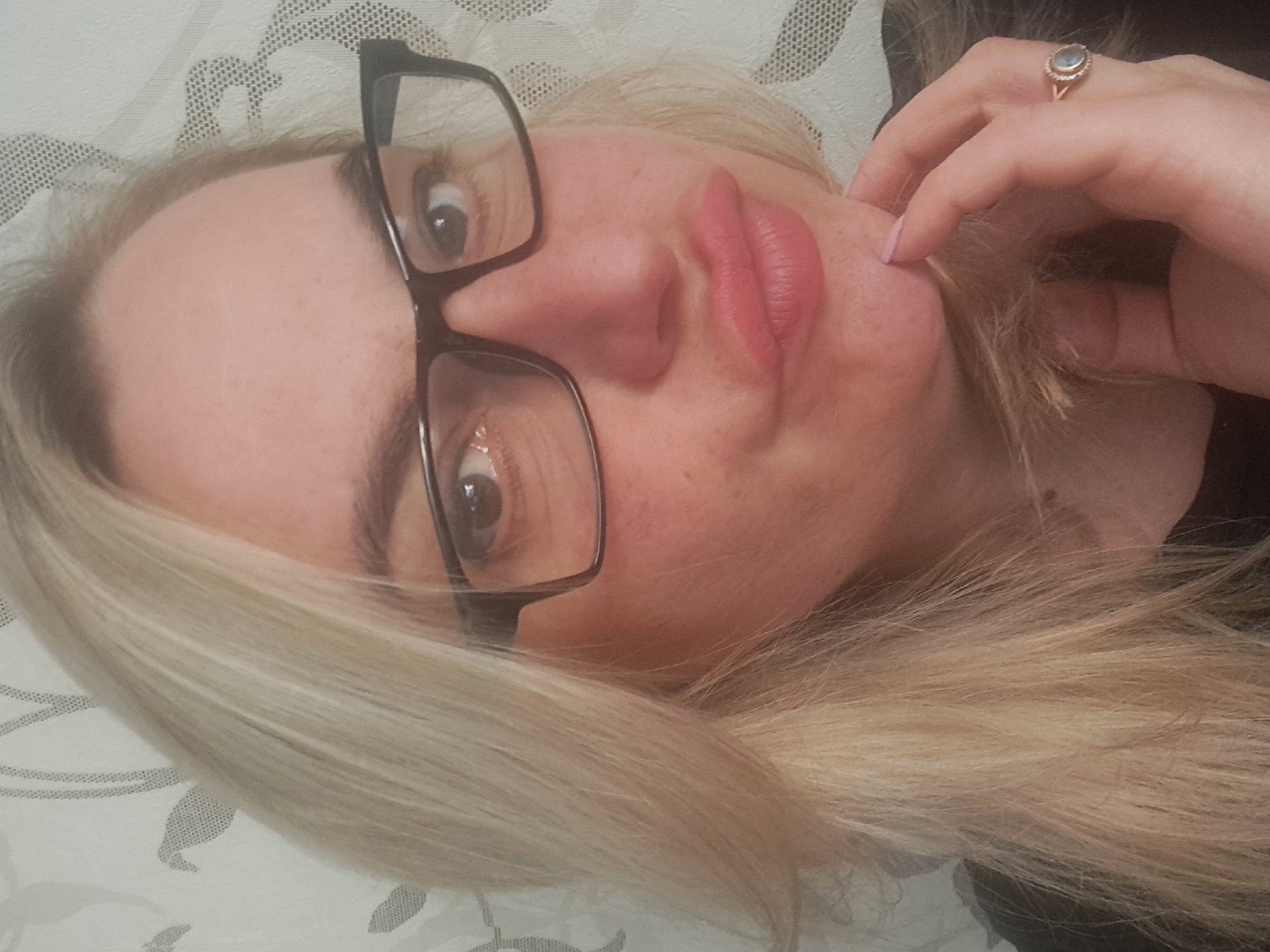 A woman with blonde hair and glasses is lying down, looking directly at the camera. Her head is tilted to the left. She is wearing a ring on her right hand, which is near her face. The background appears to be a patterned bedspread or pillow. The city of Elektrostal, Russia, cannot be ascertained from the image.