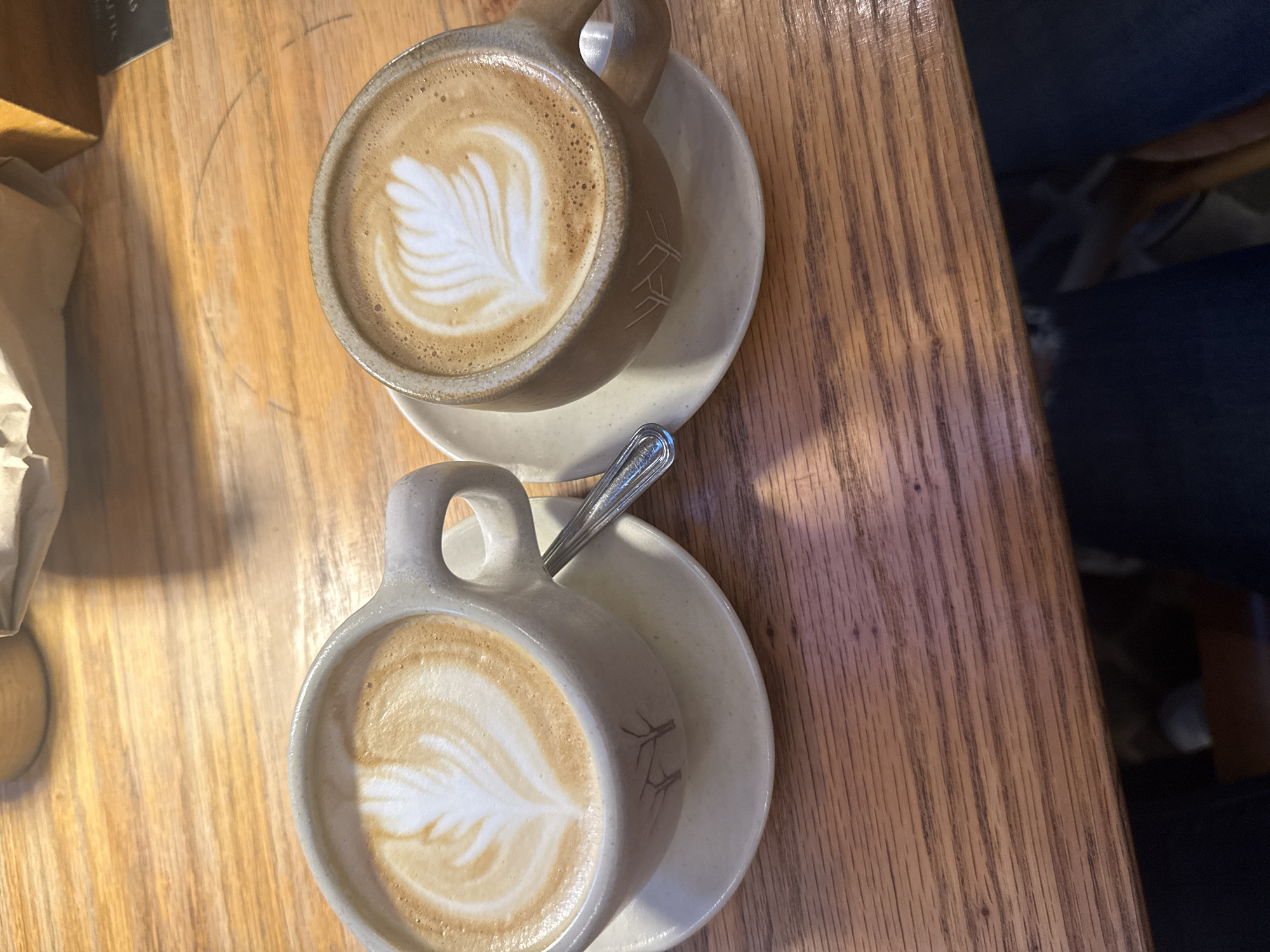 Two mugs of latte art coffee sit on a wooden table. One mug is tan and the other is light gray. Both mugs sit on matching saucers and the light gray mug has a spoon in it. A partially visible paper bag sits to the left of the mugs. A person is partially visible to the right. The location is San Cristóbal, Mexico.