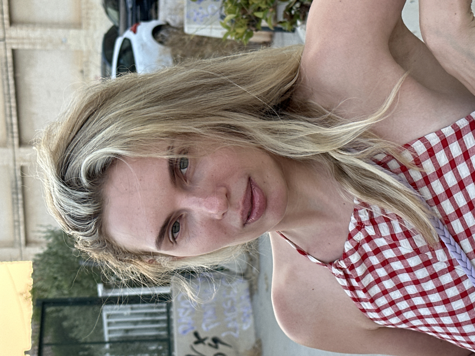 The image shows a close-up, medium shot of a woman with blonde hair and fair skin. She is wearing a red and white gingham halter top. Her hair is long and appears to be slightly windswept. The background is blurred but suggests an outdoor urban environment, possibly with a building and some vegetation visible. There are cars in the background, and graffiti is visible on a wall to the left. The lighting suggests it is daytime, possibly late afternoon due to the warm tones. The location of Benimàmet-beniferri, Spain, is not directly evident from the visual cues in the image itself.