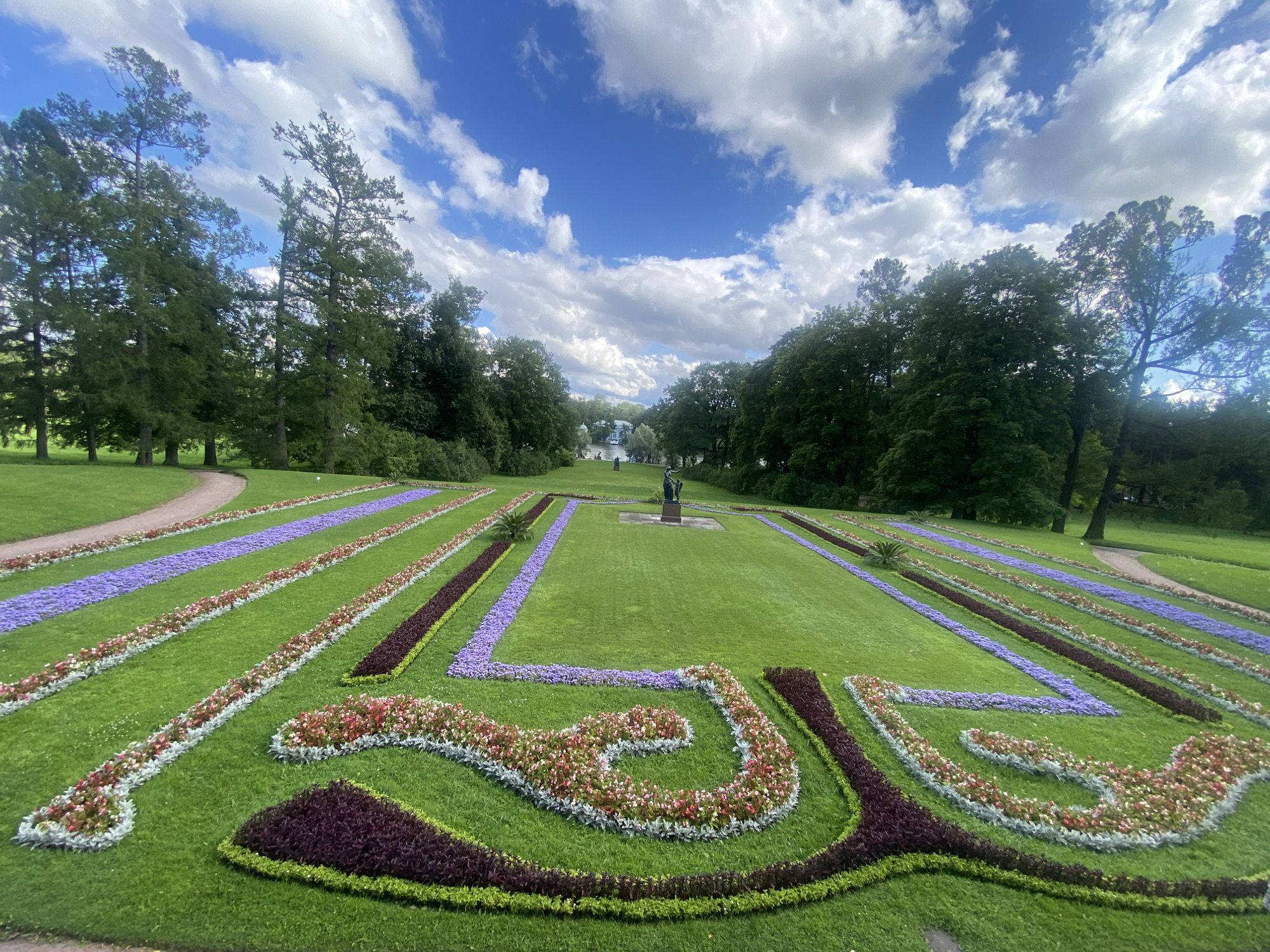 This image shows a formal garden in Pavlovsk, Russia. The foreground features a meticulously manicured lawn with intricate flowerbeds arranged in geometric patterns. The flowerbeds are composed of a variety of colorful flowers, including purple, pink, red, and white blossoms, bordered by dark green and purple foliage. In the middle ground, a statue stands on a pedestal in a large, open grassy area. Beyond this, a body of water is visible, with a white building on its far shore, and a path winding through manicured trees. The background is dominated by a dense forest of mature green trees, and the sky above is a vibrant blue with fluffy white clouds. The overall scene depicts a serene and well-maintained park landscape.