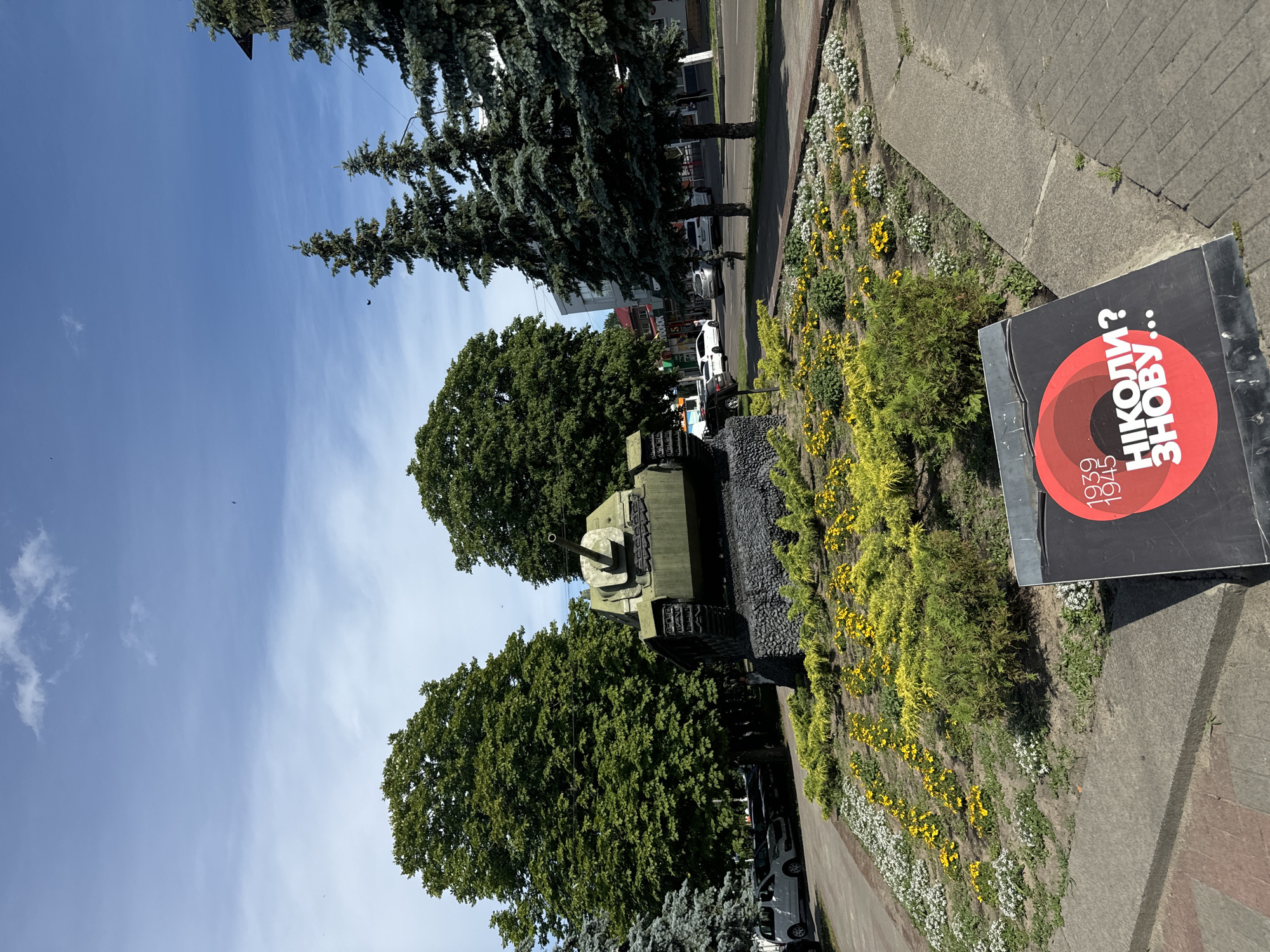 The image shows a memorial display in Sarny, Ukraine. The primary subject is a green tank, likely a historical artifact, placed on a bed of dark gravel. Surrounding the tank is a landscaped area with a variety of plants, including trees, shrubs, and flowering plants, with yellow and white flowers visible. A paved walkway runs alongside the landscaped area.
To the right of the memorial, a large black sign with a red circle is displayed. The red circle contains text in white, which appears to read "НІКОЛИ? ЗНОВУ..". Below this, smaller text reads "1939 1945". This suggests the memorial commemorates events related to World War II and serves as a reminder to prevent future conflicts.
In the background, a street with a few cars is visible, as well as buildings and tall trees. The sky is blue with some scattered clouds, indicating a daytime scene with fair weather. The orientation of the image is rotated, with the sky appearing on the left and the ground on the right. The overall impression is one of a solemn and reflective public space, likely in a park or along a prominent street in Sarny.