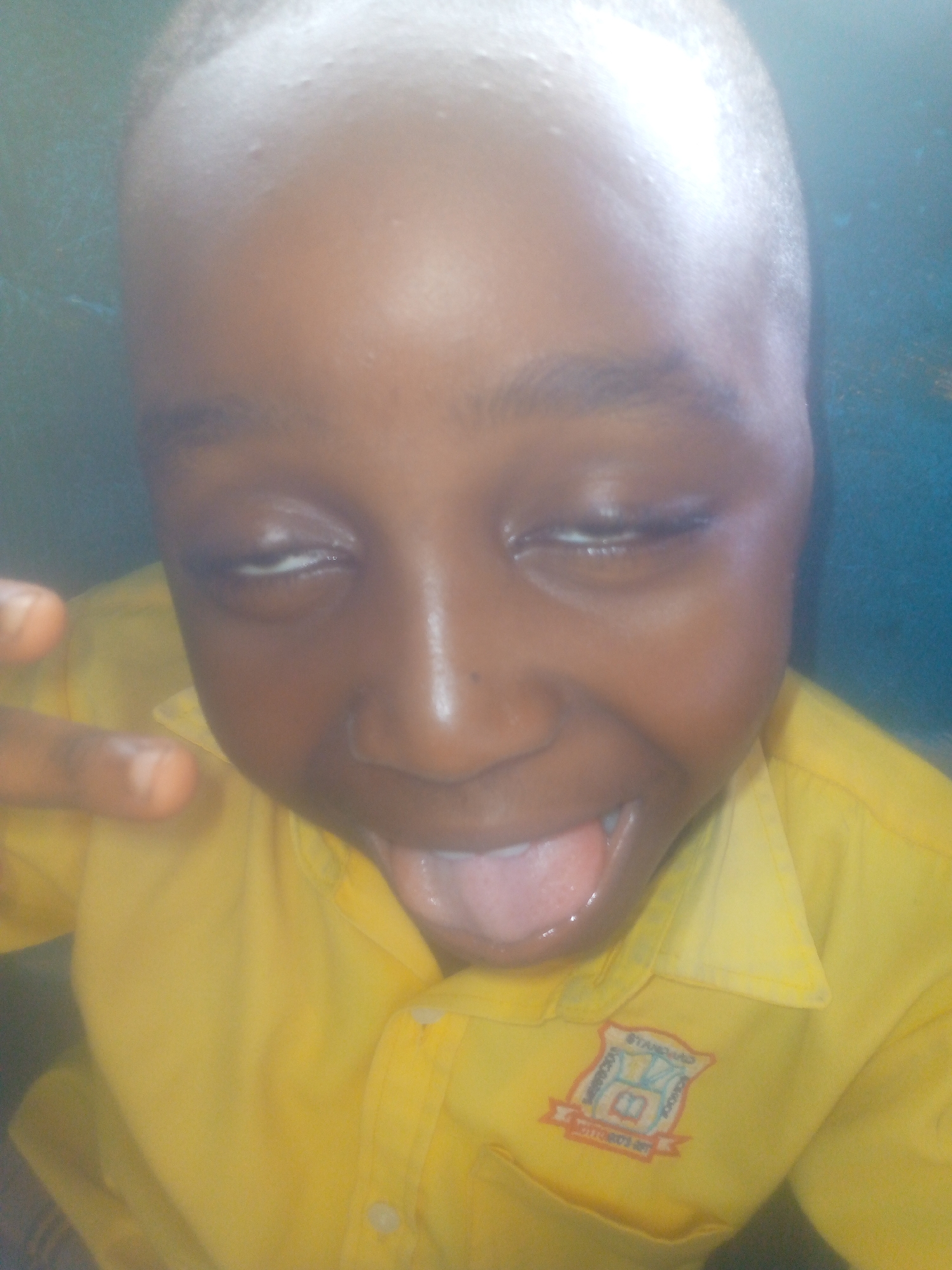 A close-up shows a young boy with dark skin, sticking his tongue out. His eyes are mostly closed, and he is wearing a yellow collared shirt with a school emblem on the left chest. The location is unidentified.