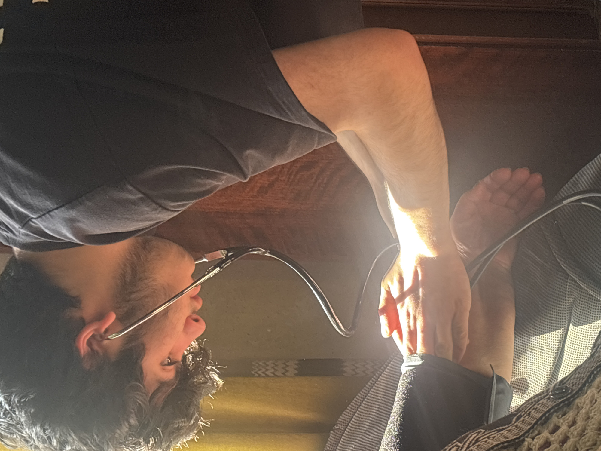 A young man with dark, curly hair, a light beard, and wire-rimmed glasses is depicted in a close-up, indoor shot. He is wearing a dark t-shirt. A stethoscope is in use, with one earpiece in his left ear and its black tubing extending towards his lower body. His right arm and hands are brightly illuminated by warm, natural light, suggesting sunlight streaming in. He appears to be holding the stethoscope's diaphragm near his shin or ankle, where a part of his bare foot is also visible, indicating a self-assessment or medical check-up. His expression is focused and contemplative.
The background includes a dark, polished wooden surface above him, possibly part of furniture like a table or bed frame. To his right, various textured fabrics are present, including a striped grey and white material and a lighter, possibly crocheted or patterned, fabric. A muted yellowish surface is visible behind his head. The strong sunlight creates a warm ambiance. No text is visible in the image.