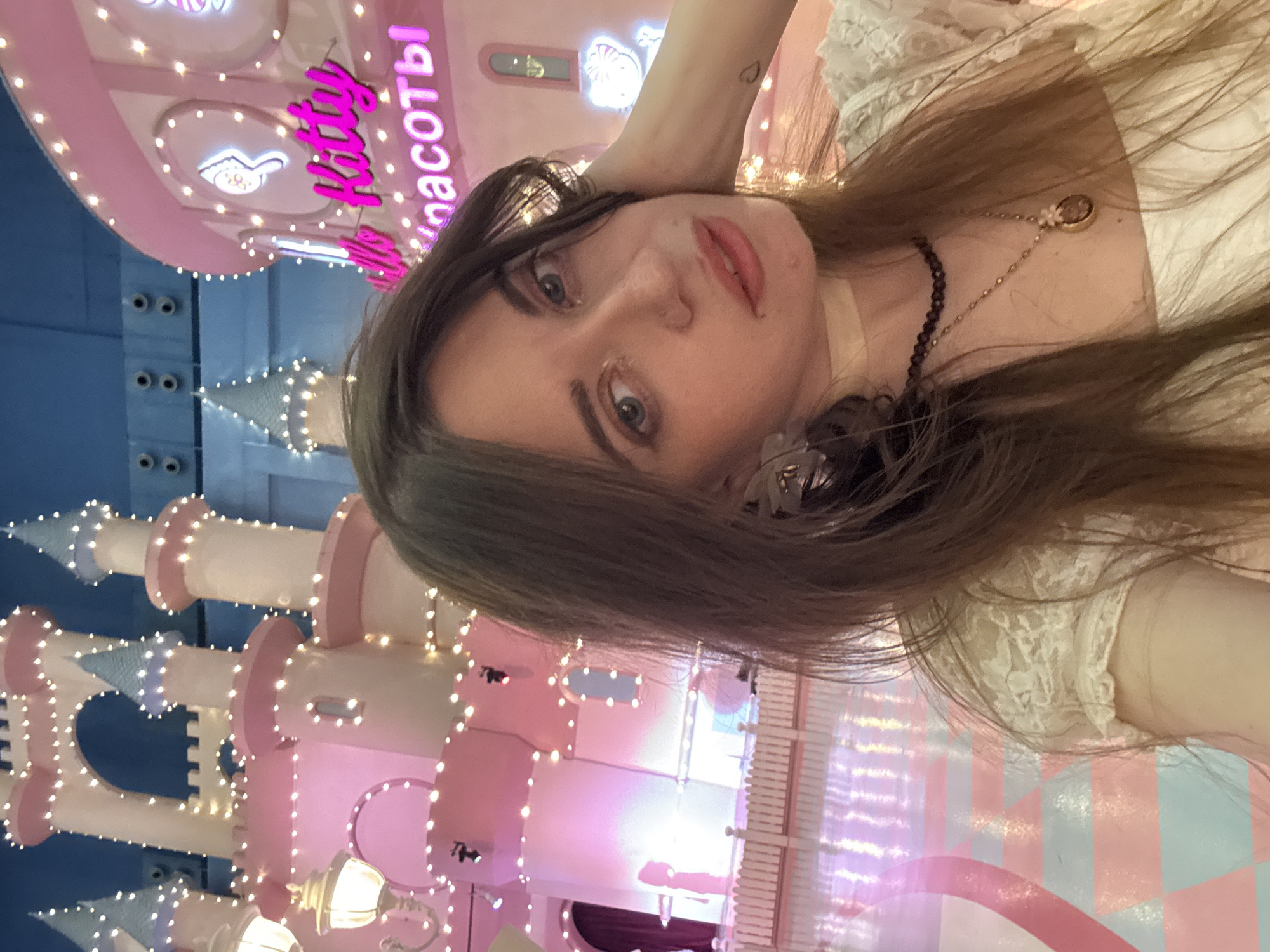 A young woman takes a selfie indoors at a Hello Kitty-themed establishment in Moscow, Russia. The background includes a pink castle-like structure adorned with lights, with signage in Russian (likely "Kitty Beauty"). The woman has long brown hair and is wearing a white lace top and necklaces. The overall lighting is soft and pink-toned.