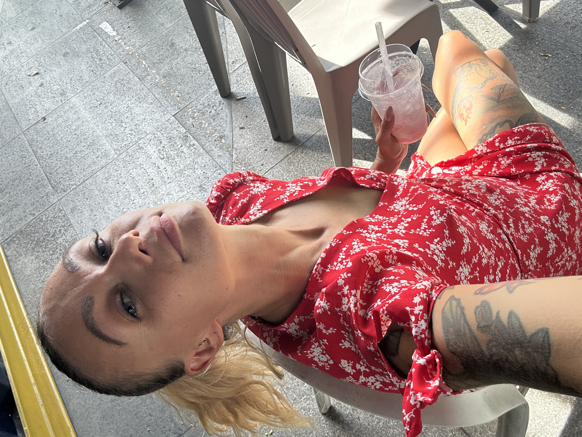 A woman with short blonde hair sits on a white plastic chair, holding a plastic cup with a straw and ice inside. She wears a red dress with a white floral print. Her left leg has a large tattoo on it. The background shows a paved area with a grey brick pattern and other white plastic chairs. The location is likely a café or outdoor seating area in a city. The image doesn't provide enough information to accurately specify the location within Đồng Nai, Vietnam.
