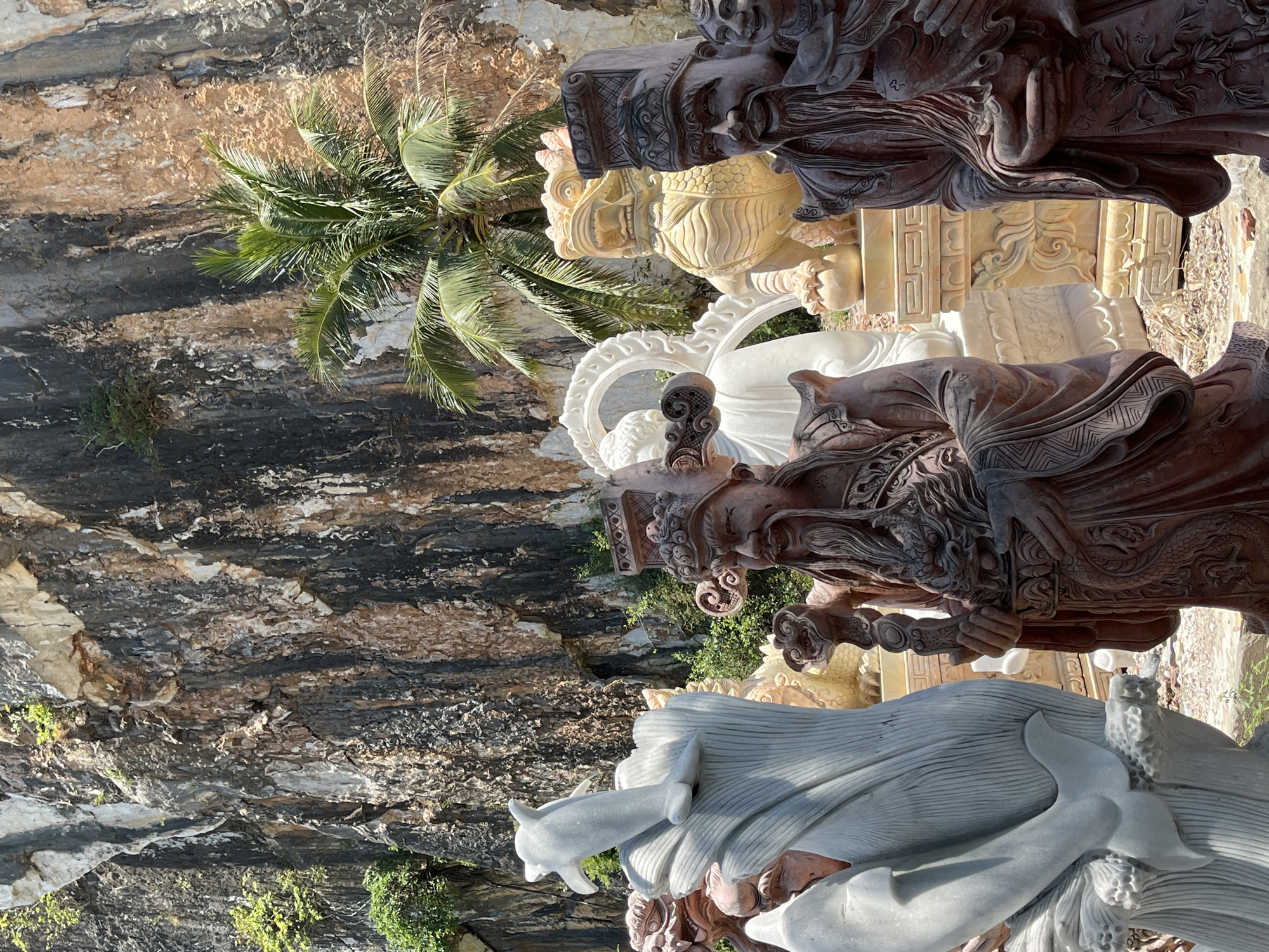 The image shows a collection of stone statues set against a cliff face and a palm tree, which is located in Đà Nẵng, Vietnam. The statues, varying in color from gray to dark brown, appear to be elaborately carved figures. The cliff face behind them is a mix of dark and light-colored rock, with some greenery growing on it. A palm tree is partially visible in the background, providing a sense of the tropical environment.