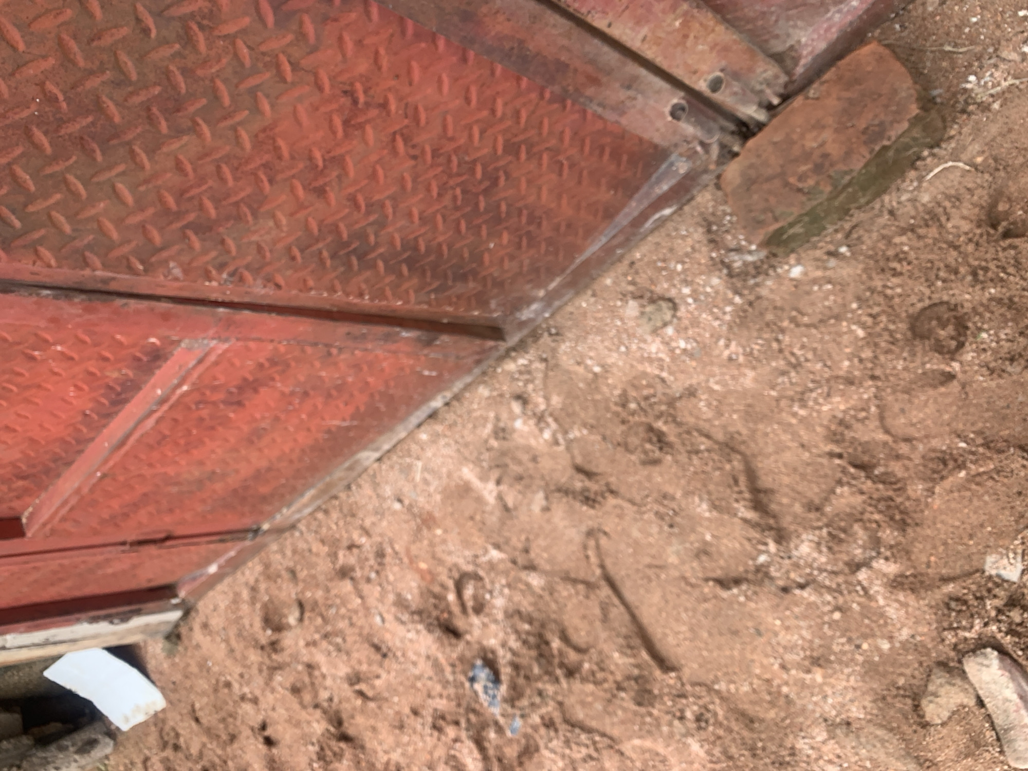 The image depicts a close-up view of a red metal door with a diamond plate pattern, positioned next to a sandy or earthy ground. A single brick lies on the ground near the door. The ground shows some indentation marks, possibly from footprints or other objects. There is also a piece of white material visible in the upper left corner. The scene appears to be outdoors, potentially in a construction or unpaved area. The location is Jauro Yinu, Nigeria.
