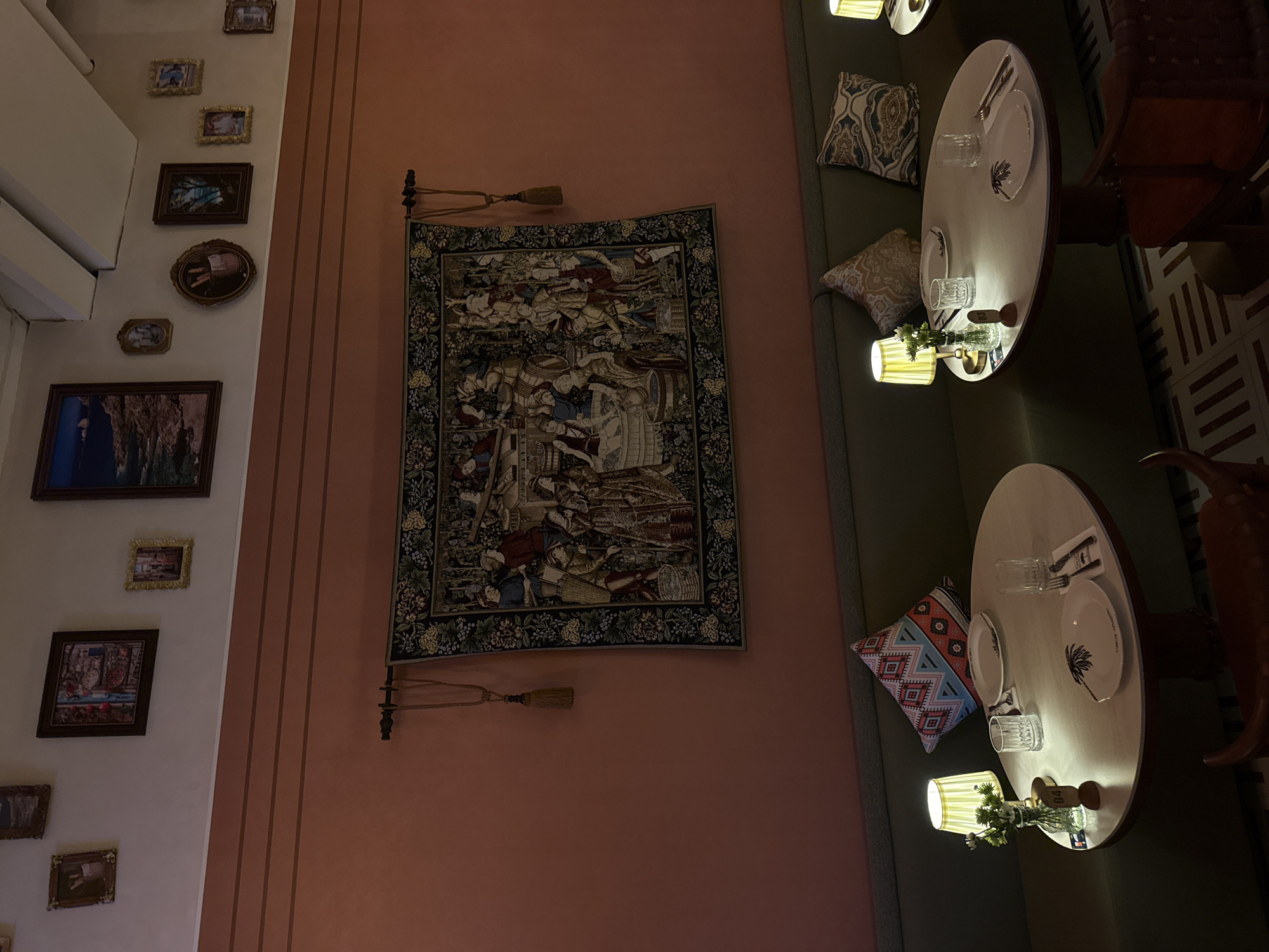 This image depicts the interior of a restaurant, likely in Yiwu, China, characterized by its warm, intimate lighting and eclectic decor. The focal point of the scene is a large, ornate tapestry hanging on a salmon-colored wall, depicting a historical or mythological scene with numerous figures. Above the tapestry, a row of framed pictures and artwork adorns the upper wall.
In the foreground, two round tables are set for dining, each with place settings including plates, glasses, and cutlery. Small lamps with pleated yellow shades cast a soft glow on each table, highlighting a vase with a few delicate flowers on the left table. Decorative pillows with geometric and paisley patterns are arranged on the banquette seating behind the tables. The floor features a tiled pattern with alternating colors. The overall ambiance suggests a cozy and aesthetically curated dining space. There are no people visible in the image.