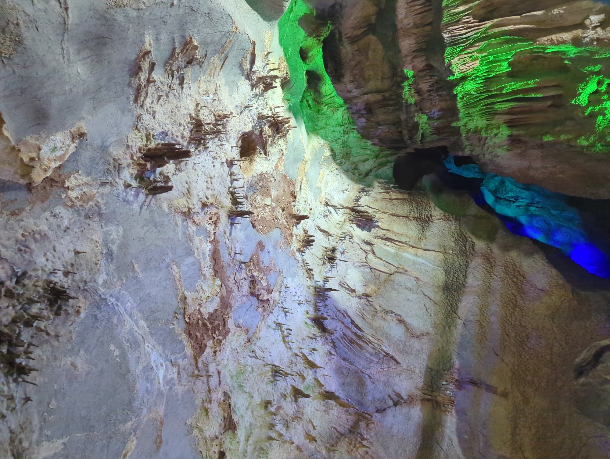 The image depicts the interior of a cave, likely Ledenika Cave, located near Vratsa, Bulgaria. The cave ceiling is adorned with numerous stalactites, varying in size and shape. The rock formations are illuminated with colored lights, predominantly green and blue, highlighting the textured surfaces and casting shadows. The cave walls exhibit layered rock strata and smaller formations resembling icicles. The lighting creates an otherworldly atmosphere, emphasizing the geological features of the cave.