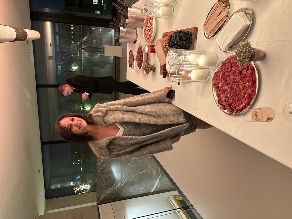 A woman in a gray fur coat stands next to a long table laden with food. The table includes platters of meat, nuts, grapes, crackers, and other appetizers, as well as candles and wine. In the background, a man in a dark suit is looking at his phone near a large window with a city view at night. The scene appears to be an upscale event or party.