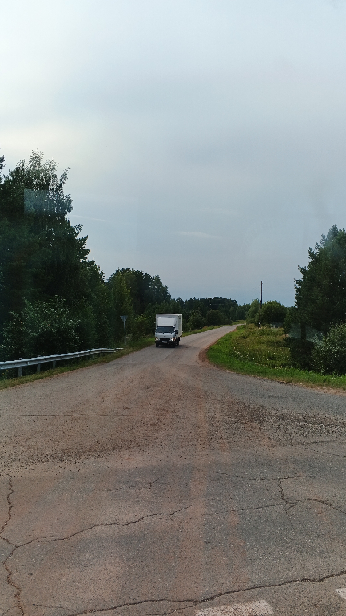 The image depicts a rural road scene, likely in or near the village of Kiyasovo, Russia, given the context. The road is asphalt and shows signs of wear, with visible cracks in the foreground. A white box truck is traveling away from the viewer on the road, which curves to the right. The road is flanked by dense green trees and foliage on both sides, with a bright, overcast sky dominating the upper portion of the frame. A metal guardrail is present on the left side of the road, and a traffic sign is visible on the left verge. The overall environment suggests a temperate climate with lush vegetation.
