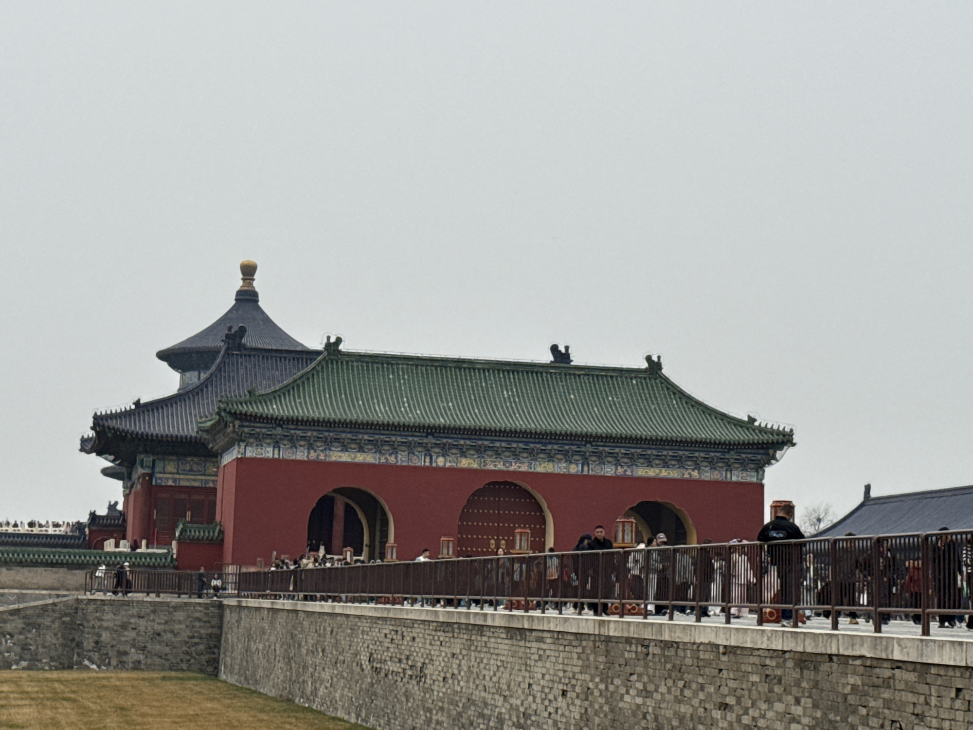 This image displays a section of the Temple of Heaven complex, situated in Dongcheng District, Beijing, China, under an overcast sky.
In the mid-ground, a raised stone walkway with a dark brown metal railing extends horizontally across the frame. Below the walkway is a textured stone retaining wall, partially obscured on the left by a patch of dry, light brown grass.
The background is dominated by traditional Chinese architecture. To the left, a multi-tiered building with a dark-tiled roof culminates in a golden finial, its eaves adorned with intricate patterns. Adjacent to it on the right, a long, red-walled structure with a green-tiled roof features multiple prominent arched gateways. Some gateways are open, revealing interior darkness, while others are closed by large, red, studded doors. The eaves of this red building are also decoratively painted. A portion of another dark-roofed building is visible to the far right background.
Multiple individuals are present on the raised walkway and near the entrances of the red-walled building. These individuals are dressed in various civilian clothing, including dark jackets and coats. People are observed walking in both directions along the walkway, standing near the railing, and gathering in groups near the arched doorways of the main structures. No specific actions beyond pedestrian movement and congregation are discernible.