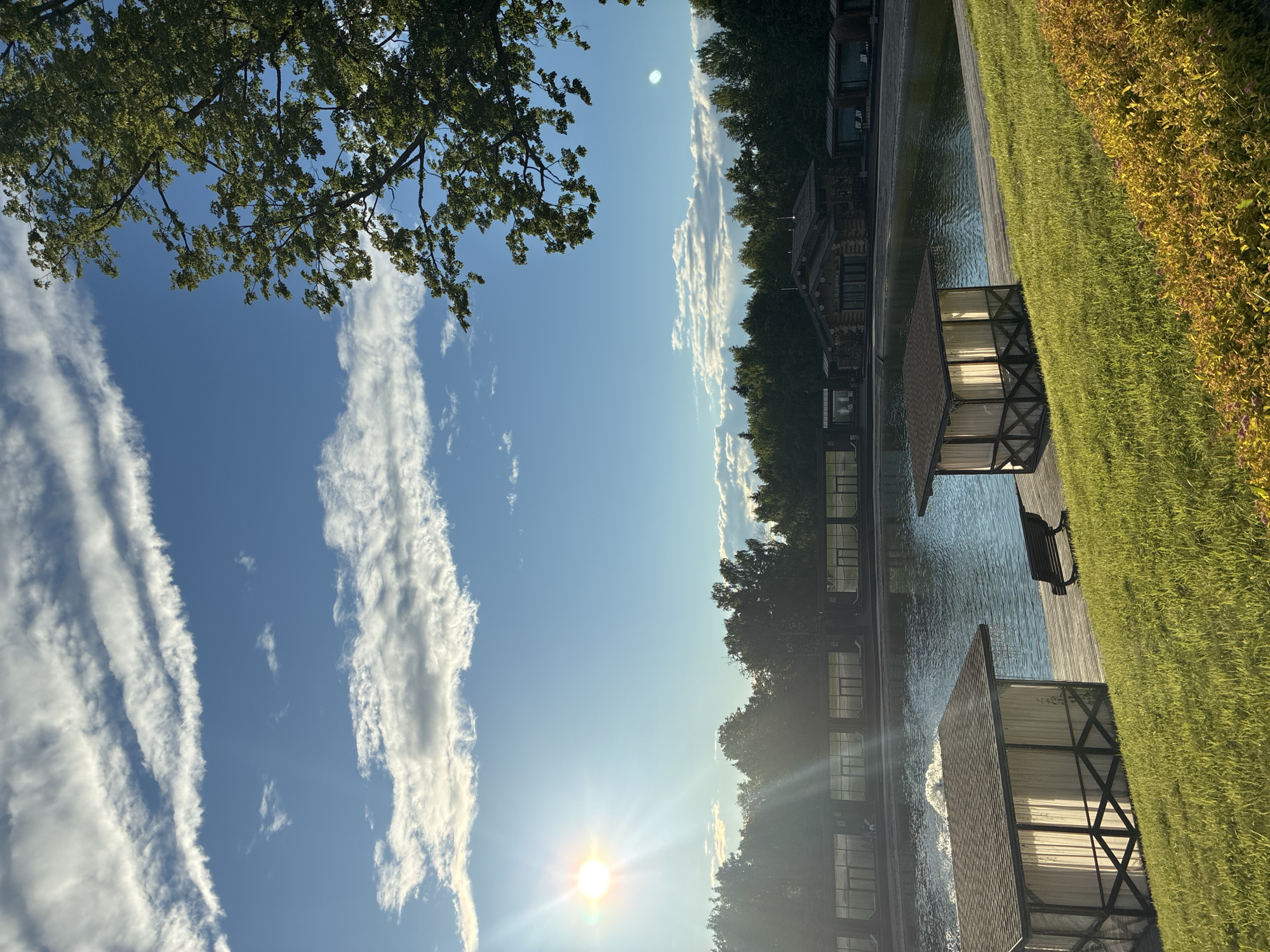 This is a landscape photograph taken outdoors, oriented vertically, with the sun low in the sky and casting a bright glare. The scene depicts a tranquil waterside setting with modern architecture.
In the foreground, a wooden deck runs alongside a calm body of water. On the deck, there are at least two structures with gabled roofs and open sides framed by dark wood beams, suggesting they might be gazebos or cabanas. One of these structures has a bench in front of it. Further into the water, more of these structures are visible, extending along the waterfront. The buildings appear to be made of dark wood and stone, with large windows.
To the right of the deck and water is a lush, green lawn that slopes upwards, bordered by vegetation with yellow and orange hues.
The sky is a clear, bright blue, with streaks of white, wispy clouds. The sun is prominently featured, creating a lens flare effect with its rays stretching across the image. The low angle of the sun suggests it is either early morning or late afternoon. The overall atmosphere is peaceful and serene, bathed in warm sunlight.
There is no visible text in the image. The location is Peresvet, Russia.