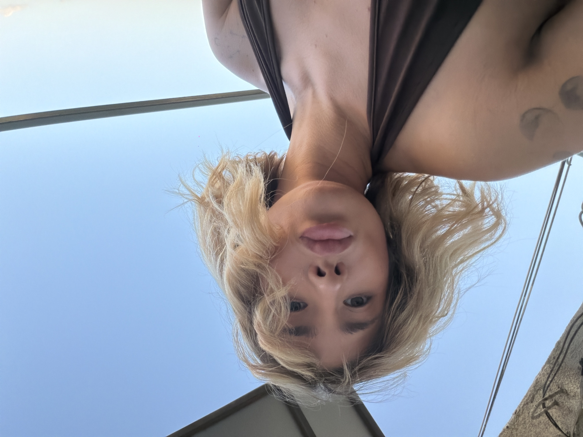 In an outdoor selfie taken from a low angle against a clear blue sky, a young woman with blonde, wavy hair and prominent lips looks directly at the camera. She is wearing a brown halter-neck top. Her left shoulder and upper arm are visible, with a faint tattoo of three rounded shapes on her arm. A portion of a light-colored, slatted overhang and a dark railing are visible in the background. The image does not contain any elements that suggest a specific location in Tbilisi, Georgia.