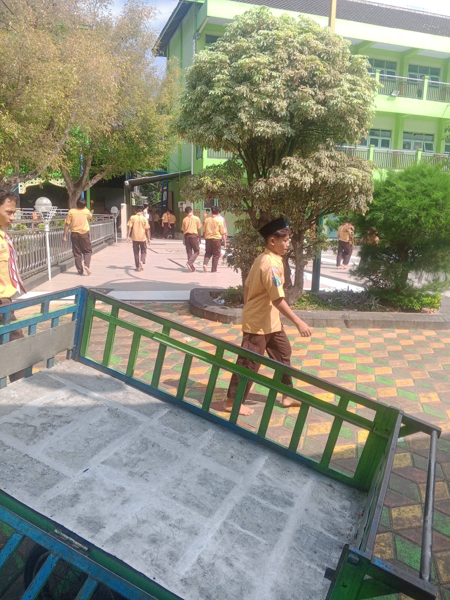 A group of Indonesian students in brown uniforms and scout scarves walk on a patterned tiled walkway in front of a light green, multi-story building with balconies. Some students are walking towards the camera, while others are walking away. The building appears to be a school. In the foreground, a green metal railing is partially visible. The environment is sunny with trees and some vegetation. The specific location is in Ponorogo, Indonesia.