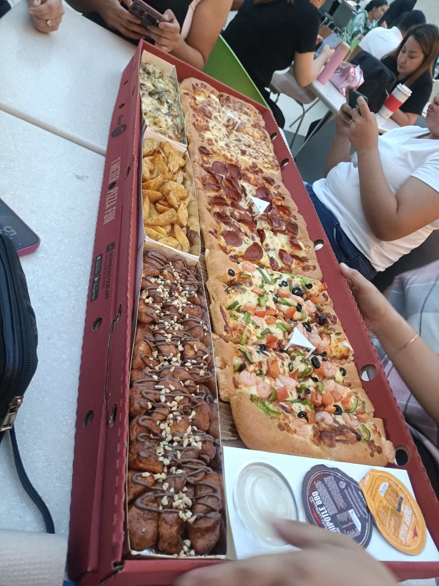 The image depicts a close-up, elevated view of a large, rectangular pizza box filled with various food items. The pizza box is red and has text that reads "1 METER PIZZA FUN" on the side. Inside the box, from top to bottom, are: a section of a creamy white dish with visible green ingredients, a section of golden potato wedges, two large rectangular pizzas with different toppings (one with pepperoni, the other with shrimp, peppers, and olives), and a section filled with chocolate-covered bread rolls topped with chopped nuts and chocolate drizzle. To the right of the pizzas, three small condiment containers are visible. In the background, several people are seated at tables, and a hand is visible in the foreground reaching towards the food. The overall impression is a casual gathering centered around food. The image does not provide specific visual cues to definitively pinpoint its location in Dubai, United Arab Emirates.