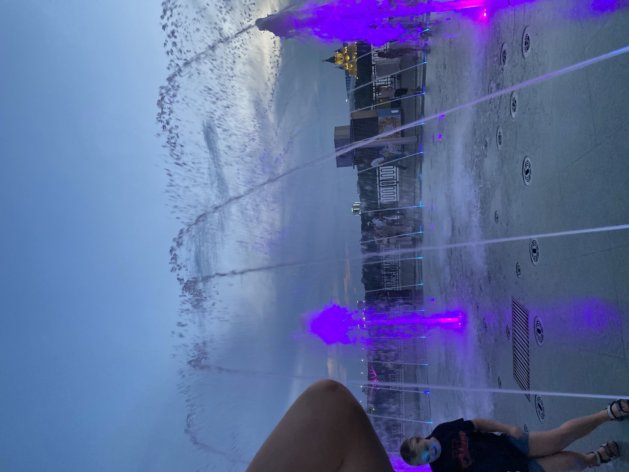 This image captures a vibrant evening scene at a public fountain in Nizhny Novgorod, Russia. The fountain features numerous jets of water illuminated with bright purple and blue lights, creating a dazzling display against the dusky sky. In the foreground, a young girl is seen walking, her face partially obscured by a spray of water. She is wearing a dark t-shirt and denim shorts, with sandals on her feet. Behind her, several other individuals are present, some standing and observing the fountain, while others appear to be walking along a promenade. The background reveals the city's skyline, including buildings and a distinct, illuminated golden domed structure, which is likely the Kremlin of Nizhny Novgorod. The ground around the fountain is wet, reflecting the colorful lights. The overall atmosphere suggests a lively public space, enjoyed by both residents and visitors during the twilight hours.
