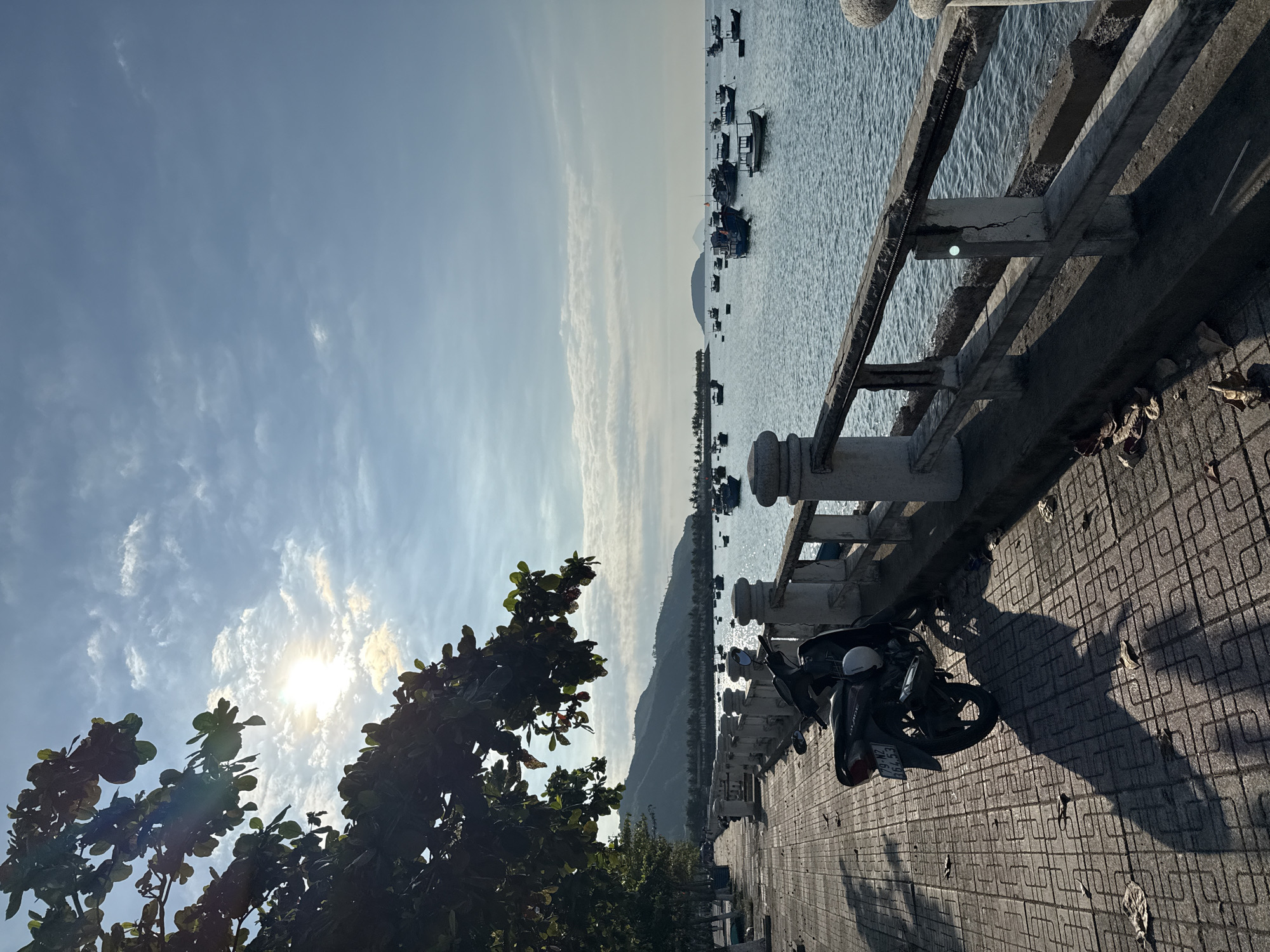 The image captures a serene seaside scene in Bac Nha Trang Ward, Vietnam. In the foreground, a dark-colored motorcycle is parked on a tiled walkway, casting a long shadow. The walkway runs alongside a concrete barrier with a wooden railing, separating it from the water. Beyond the barrier, the sea is filled with numerous small fishing boats of various colors, bobbing gently on the calm water. In the distance, a lush green mountain range forms the backdrop, with a hazy sky overhead. The sun is bright, peeking through scattered clouds, and casting a warm glow on the scene. The overall impression is one of tranquility and coastal life.
