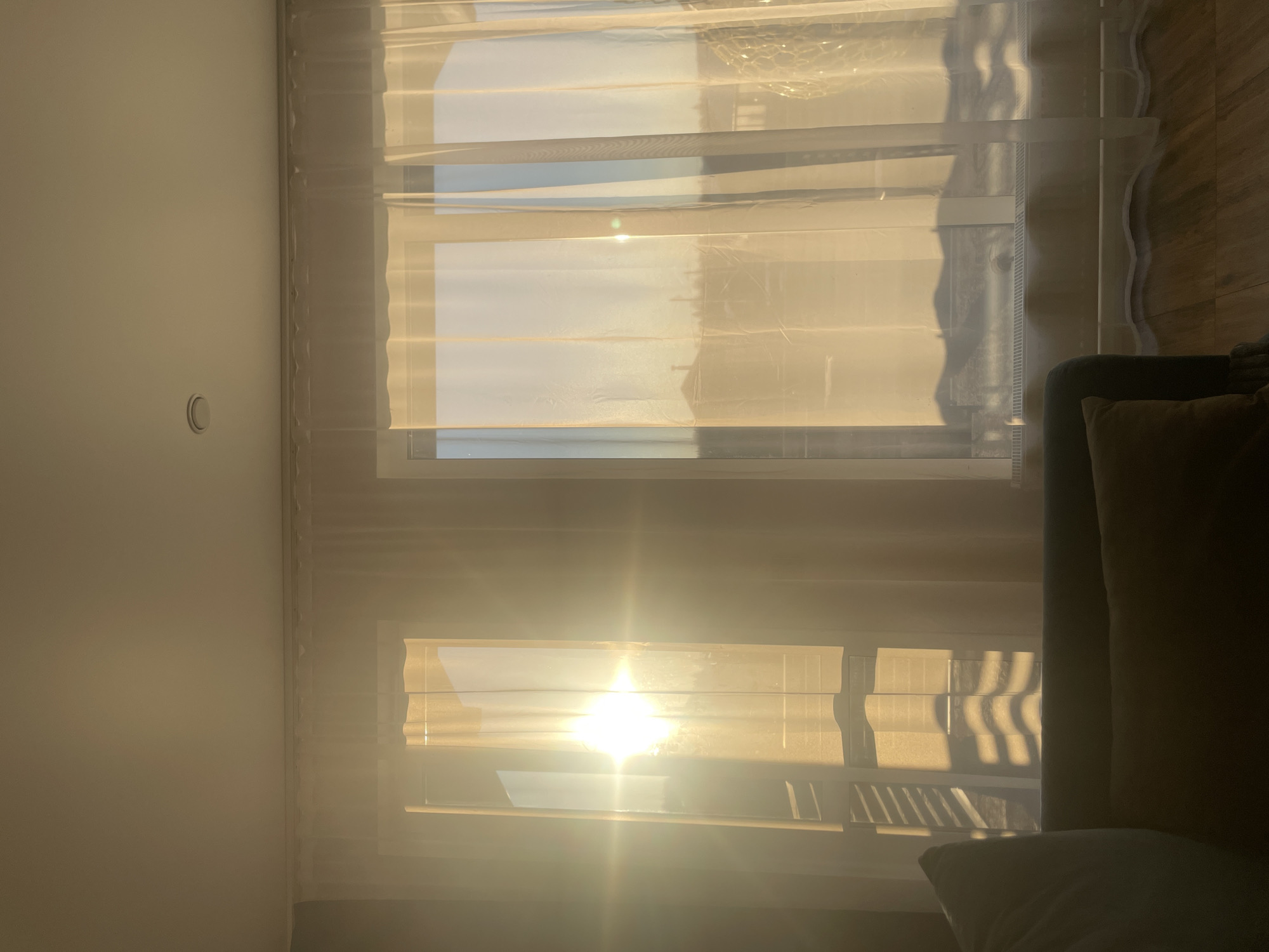 The image shows an interior view of a room in Подшивалово, Russia, with sunlight streaming through a window covered by sheer curtains and blinds. The strong light casts bright illumination and shadows throughout the room. A section of a dark-colored sofa is visible in the lower right corner of the frame. A light switch is present on the left wall. The window has two panes, which are almost completely covered by the sunlight.