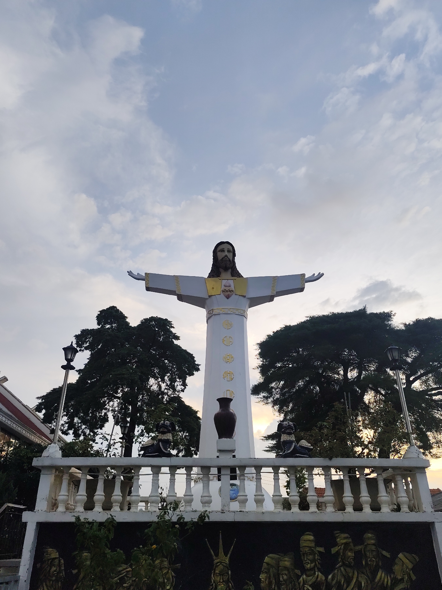 A statue of Jesus Christ, arms outstretched, stands tall against a cloudy sky. The statue is white with gold accents and is positioned on a raised platform with a white balustrade. Two dark statues, possibly guardians, flank the statue of Jesus. A dark vase is placed to the right of Jesus's feet. Below the balustrade, a mural depicts a row of figures in dark clothing and hats. Large trees with dark green foliage surround the statue. A lamppost is visible to the left of the statue. The scene appears to be outdoors in a park or religious site, possibly at dusk given the lighting. The location is identified as Lâm Viên - Đà Lạt, Vietnam.