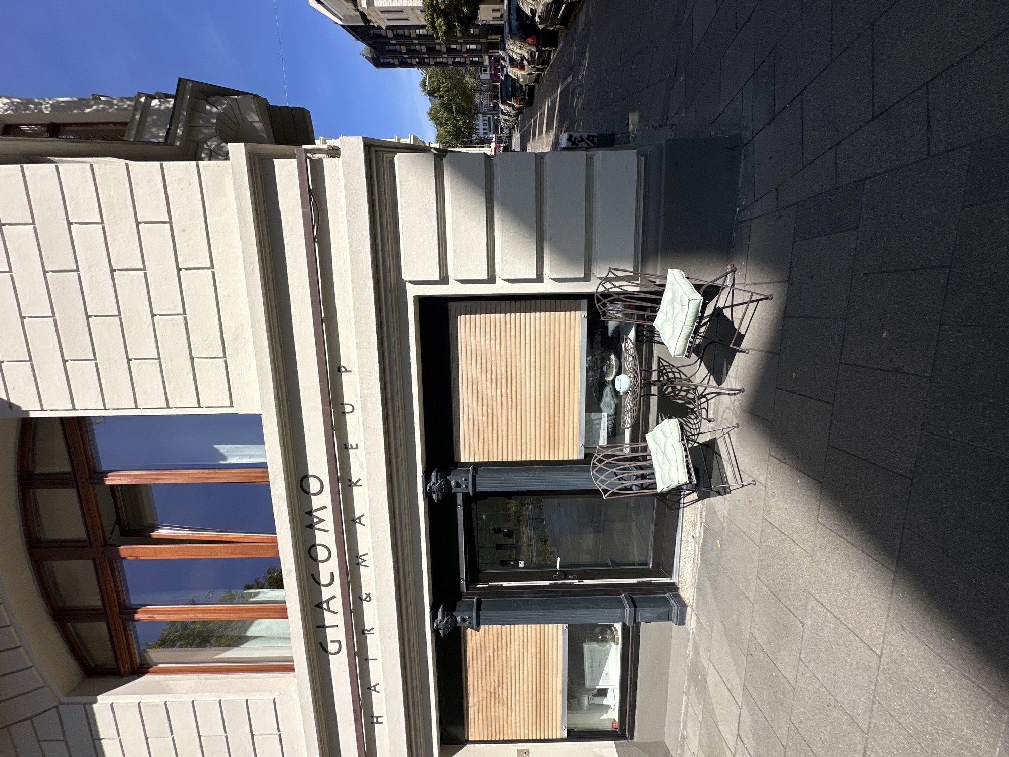 This is a slightly elevated, eye-level, full-shot, outdoor, daylight, vertical photograph of a storefront and sidewalk on a sunny day. The storefront is for a business named "GIACOMO HAIR & MAKEUP". The building has a light-colored brick facade with dark window frames. Above the storefront is a large arched window with dark wooden trim. The storefront itself has two large windows with closed wooden blinds, a dark-colored door in the center, and decorative dark pillars on either side of the door. In front of the storefront, on the gray paved sidewalk, is a small round metal table with two matching chairs. The chairs have light green cushions. A shadow from a tree falls across the sidewalk, obscuring part of the ground. Beyond the storefront and sidewalk is a street with parked cars. The street and cars appear to be in an urban setting, likely in Cologne, Germany, given the architectural style and the presence of a street with parked cars and buildings on the opposite side. The sky is clear and blue. There are no people visible in the photograph.