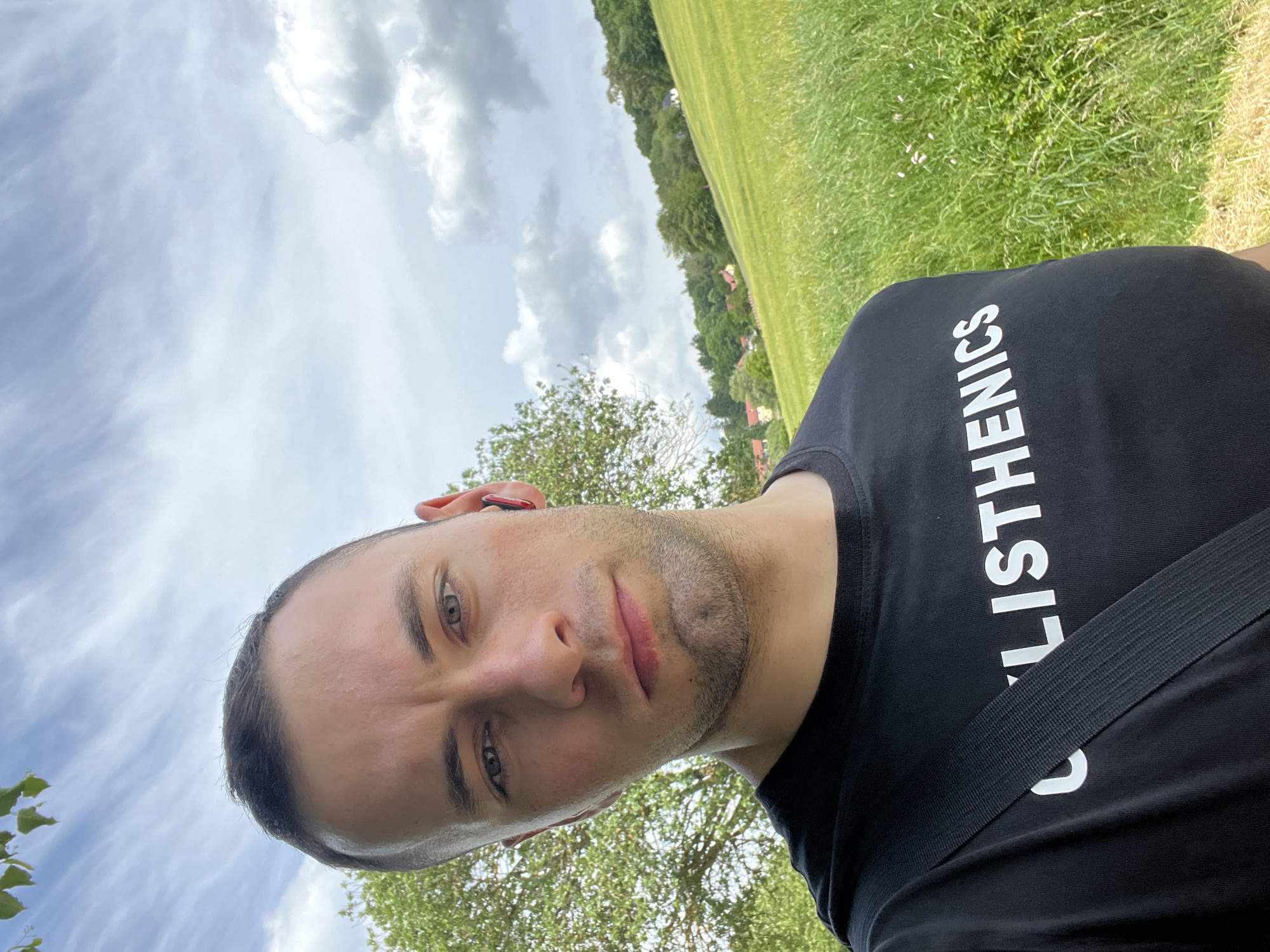 A close-up outdoor selfie depicts a man looking directly at the camera with a neutral to slightly serious expression. His head is slightly tilted, revealing short, dark hair and light-colored eyes, possibly hazel or green. He has light stubble on his chin and cheeks. A red or orange in-ear headphone is visible in his right ear. He wears a black t-shirt, across the front of which white capital letters spell out "CALISTHENICS"; the visible portion clearly reads "LISTHENICS". A black strap, possibly from a bag or seatbelt, runs diagonally across his chest.
The background on the left and upper parts of the image is dominated by a bright, cloudy sky with wispy and fuller clouds. To the right and behind the man, a vibrant green field or grassy area stretches out, bordered by a line of trees. Hints of distant structures or houses can be seen beyond the foliage. The scene is illuminated by natural daylight, indicating it is daytime. The setting appears to be an open outdoor space, perhaps a park or rural landscape.