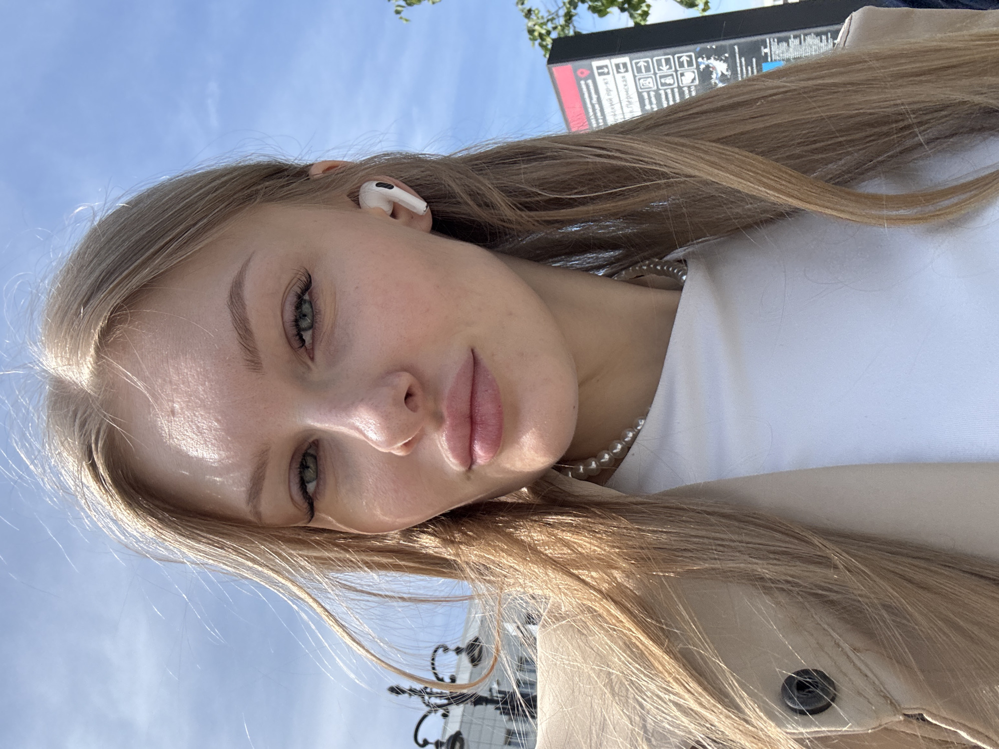 A close-up portrait of a young woman is captured outdoors on a sunny day. Her head is positioned on the left side of the frame, and she looks slightly to her right and upwards, directly at the camera, with a neutral and serene expression. She has light skin, long, straight, light blonde or ash blonde hair, and striking light-colored eyes (likely green or blue). Her lips are naturally full and slightly parted.
She wears a white wireless earbud in her right ear and a pearl necklace, visible over a white top. A light beige jacket or coat is also worn. Strong natural light illuminates her face and hair, creating bright highlights.
In the background, a clear blue sky is visible above her head. To the upper right, behind her shoulder, is a dark sign or information board featuring white text and directional arrows. Partially visible Cyrillic text includes phrases like "ский про-кт," "М. ПАРК КУЛЬТУРЫ" (referring to Park Kultury Metro Station), and "ул. Пречистенка" (Prechistenka Street). These details strongly indicate the location is Moscow, Russia, rather than Perm. Above the sign, some green tree leaves are visible. Below the sign and behind her, there's a glimpse of dark, ornate metalwork, possibly part of a fence or street furniture.
The image is presented with an unusual orientation, where the woman's head appears on the left side of the frame.
