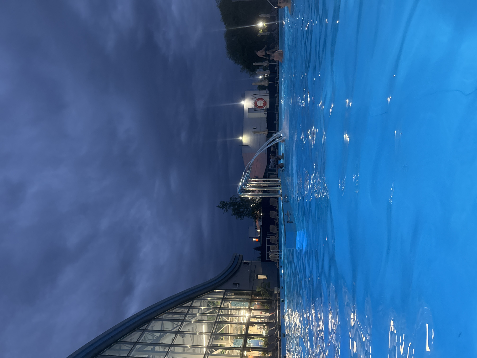 The image depicts a swimming pool at night. The pool water is illuminated with a bright blue glow. In the background, there are people swimming in the pool. There is a structure with a curved glass roof to the left of the image. The sky is dark and cloudy. In the background, further to the right, there is a building with a life preserver hanging on it, and there are a few illuminated lights visible. The location is likely in the city of Ulyanovsk, Russia.