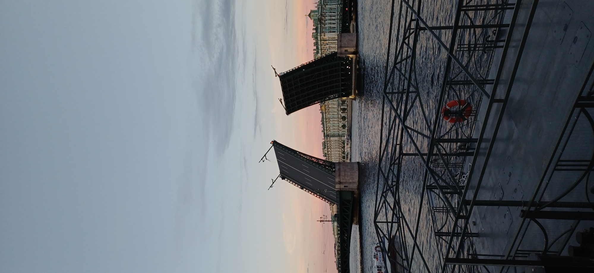 The image captures a drawbridge, likely the Palace Bridge, in Saint Petersburg, Russia, during twilight. The bridge is raised, allowing passage for a vessel. The sky is a soft gradient of pale blue to pink, with wispy clouds. The Neva River flows below, reflecting the muted colors of the sky and the bridge's structure. On the far bank, a grand, ornate building, possibly the Winter Palace, is visible, its facade illuminated by soft lights. In the foreground, a dark metal walkway or pier with railings and a life preserver can be seen. The overall scene evokes a sense of calm and the distinct atmosphere of Saint Petersburg's White Nights.