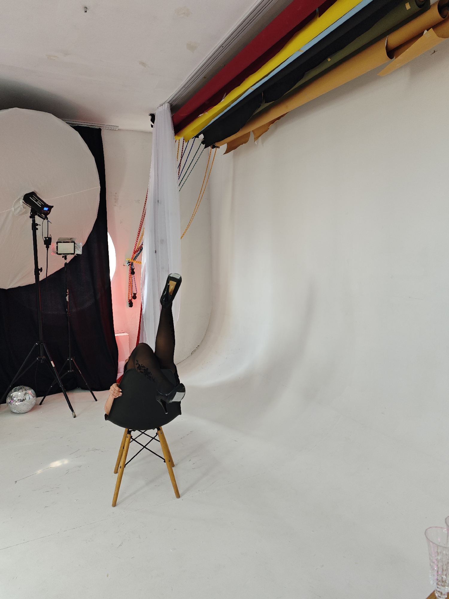 This image depicts an indoor photography studio located in Chaikovsky, Russia. The central subject is an individual positioned on a modern black chair with light wooden legs, largely obscured by the chair. Their legs are extended vertically upwards, clad in black sheer hosiery with a floral pattern on the thighs, and fitted with black high-heeled shoes featuring a gold-colored sole or heel. A bare arm and hand are visible, gripping the upper edge of the chair's back.
The studio features a continuous white seamless cyclorama acting as both floor and backdrop. On the left, professional photographic lighting equipment is present: a large white parabolic softbox mounted on a stand, accompanied by a smaller LED panel on another stand. A silver disco ball rests on the floor in the lower-left. Above the right side of the frame, several rolls of colored backdrops (red, yellow, light blue, black, olive green, and tan/light brown) are suspended from the ceiling. Adjacent to these, white sheer fabric hangs vertically, partially obscuring red and multi-colored ropes or cables adorned with small, round elements, from which a reddish glow emanates. The white ceiling shows minor stains and irregularities. In the bottom right corner, two clear, fluted glasses with a pinkish tint are partially visible. The scene suggests a professional photographic session in progress.