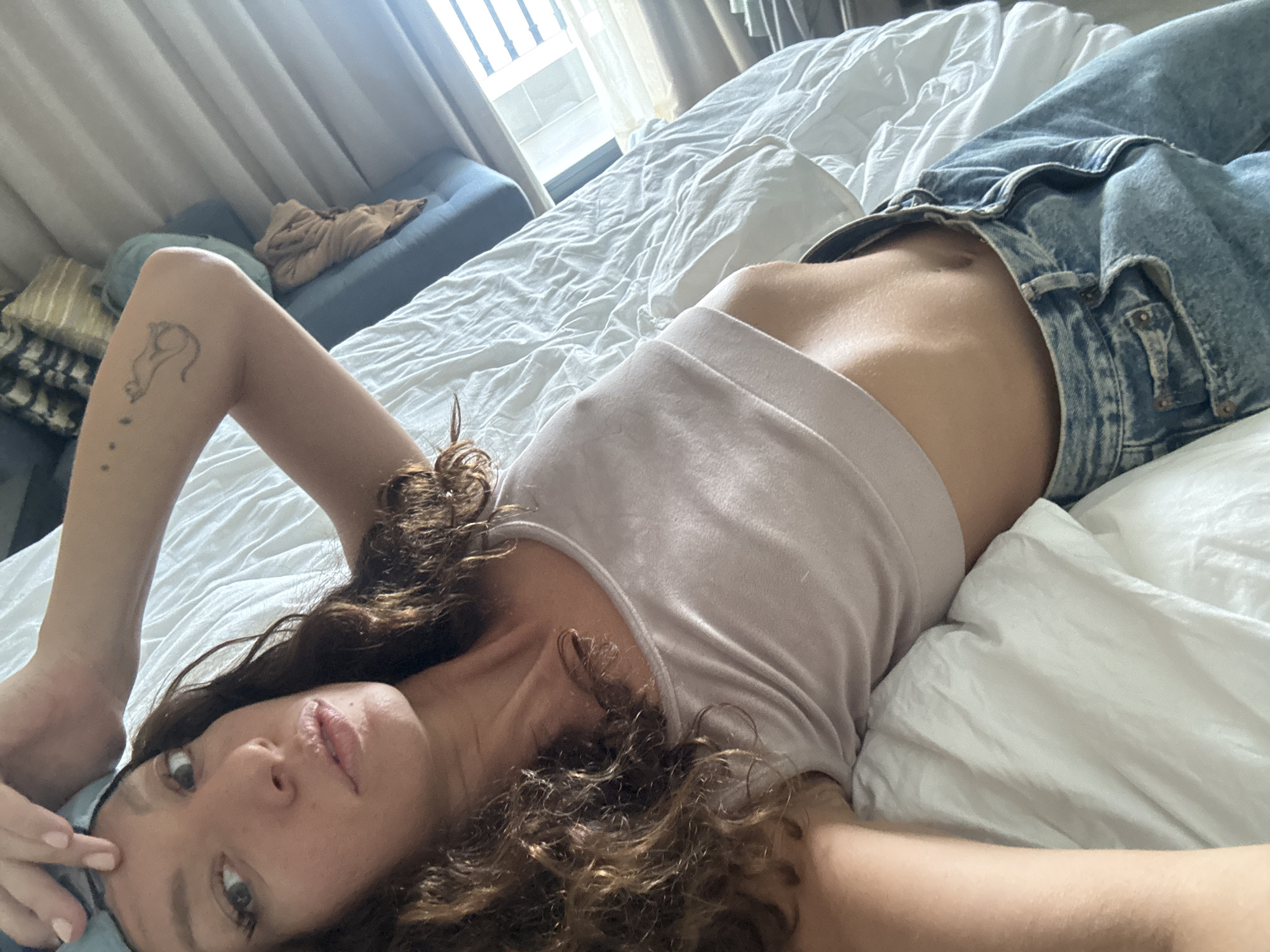 A woman is lying on a bed, wearing a light pink tank top and blue jeans. She has curly brown hair and is looking at the camera with her head tilted. Her left arm is raised, with a tattoo of a cat on her upper arm. The bed has white sheets and a pillow. In the background, there's a window with curtains, a blue couch, and some pillows. The location is not discernible from the image.