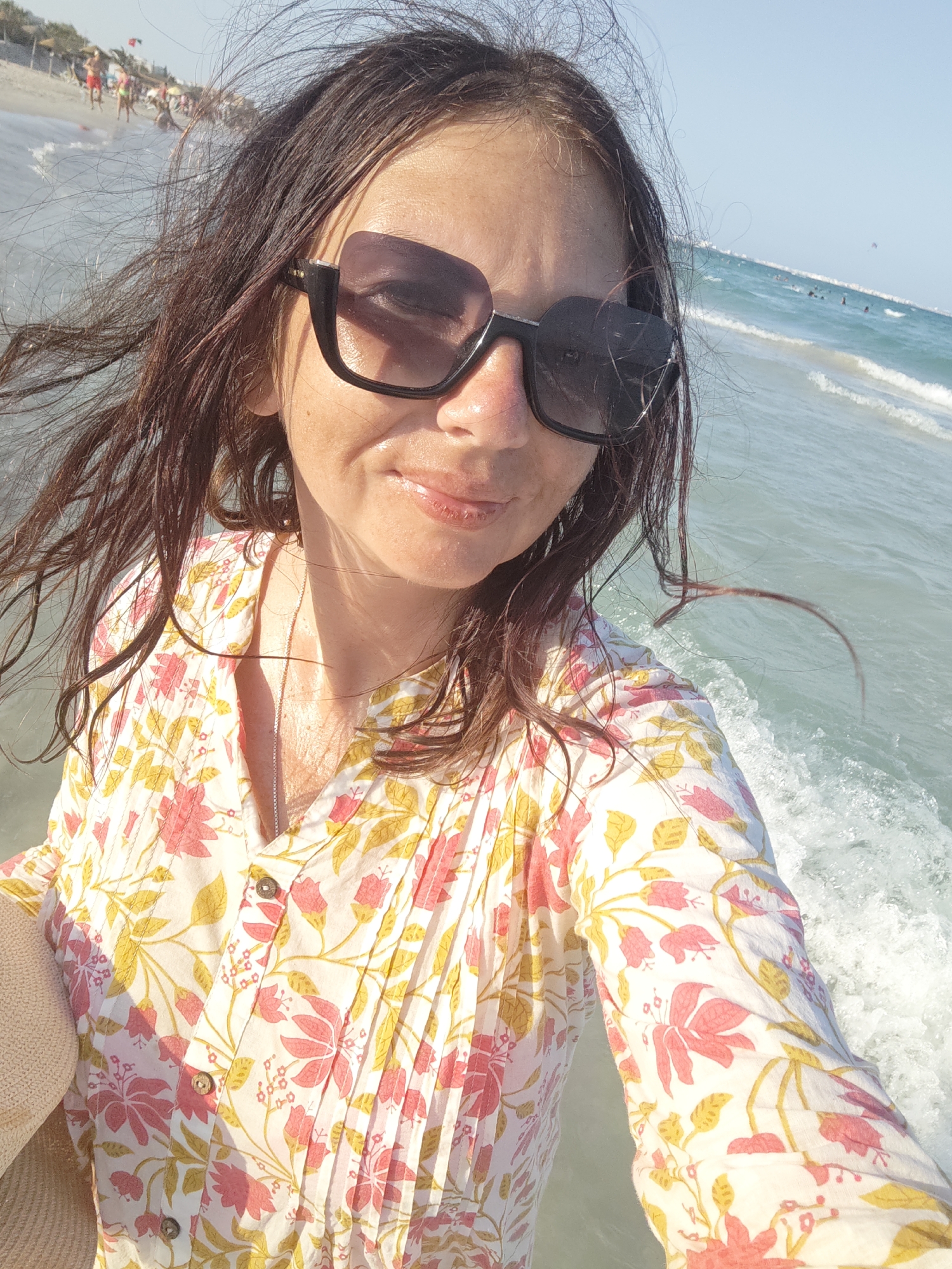 The image is a selfie taken by a woman at the beach in Mahdia, Tunisia. She is wearing a floral print top and sunglasses. The ocean is visible in the background, with waves and other beachgoers. The sun is shining, creating a bright and warm atmosphere.