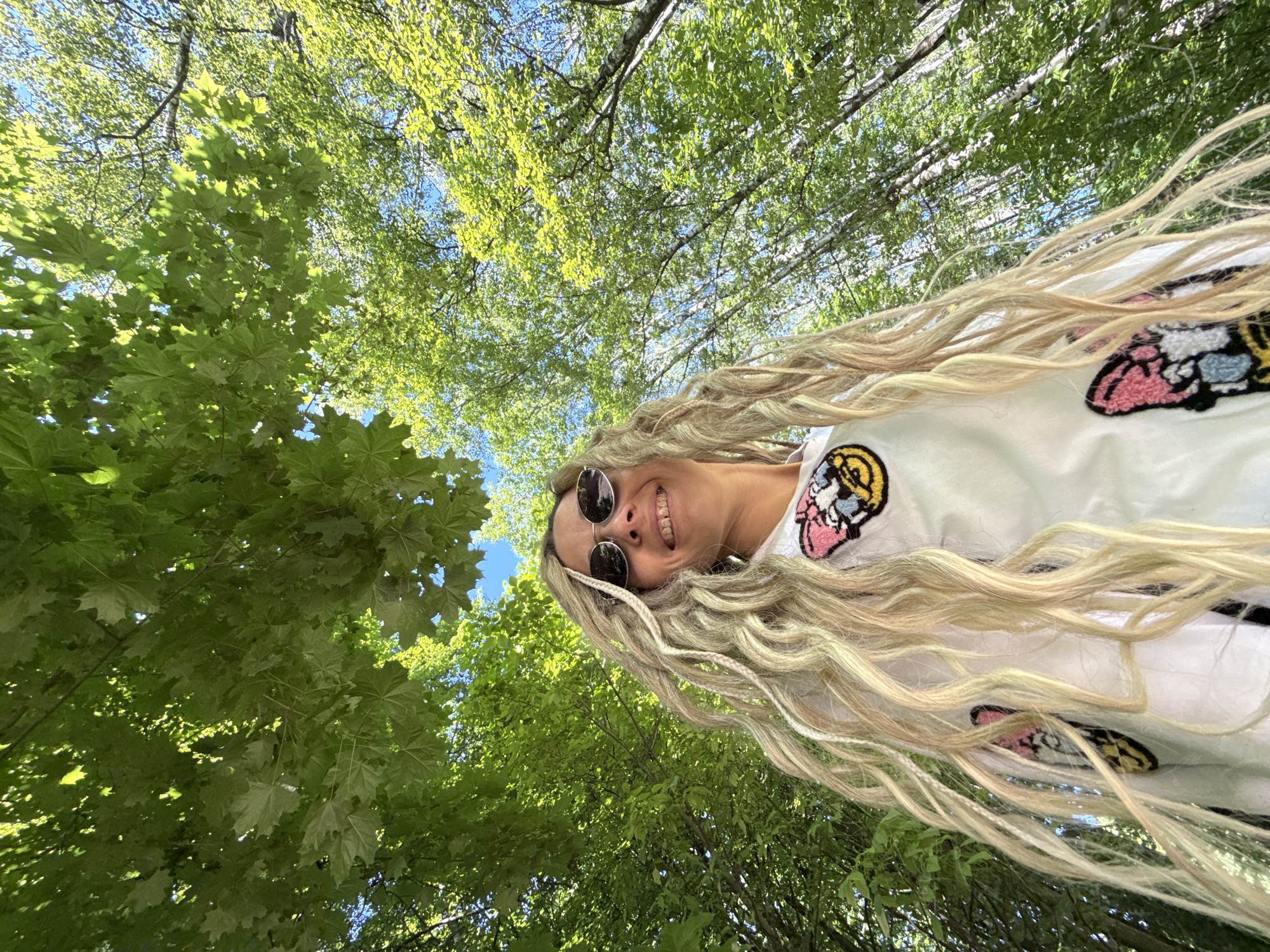 A low-angle outdoor portrait features a smiling woman positioned against a dense backdrop of lush green foliage. The woman has long, wavy blonde hair with several integrated lighter blonde braids or faux dreadlocks. She wears dark, oval-shaped sunglasses and a white long-sleeved shirt or jacket adorned with colorful embroidered patches. A prominent patch on her upper left chest displays a pink and white design, possibly a cartoon character or butterfly, outlined in black. Another patch with yellow and black elements is partially visible lower on her torso. Her head is tilted slightly back, and she is looking upwards, directly at the camera, with a wide smile revealing her teeth. The background is dominated by a vibrant tree canopy, consisting of various deciduous trees with full, bright green leaves, characteristic of spring or summer. Patches of clear blue sky are visible through the gaps in the foliage. Some trees in the upper left exhibit large, lobed leaves, while trees on the right feature slender, lighter-colored trunks and smaller leaves, indicative of birch trees. The scene is brightly illuminated by natural sunlight filtering through the leaves. The location is Rusanovo, Russia.