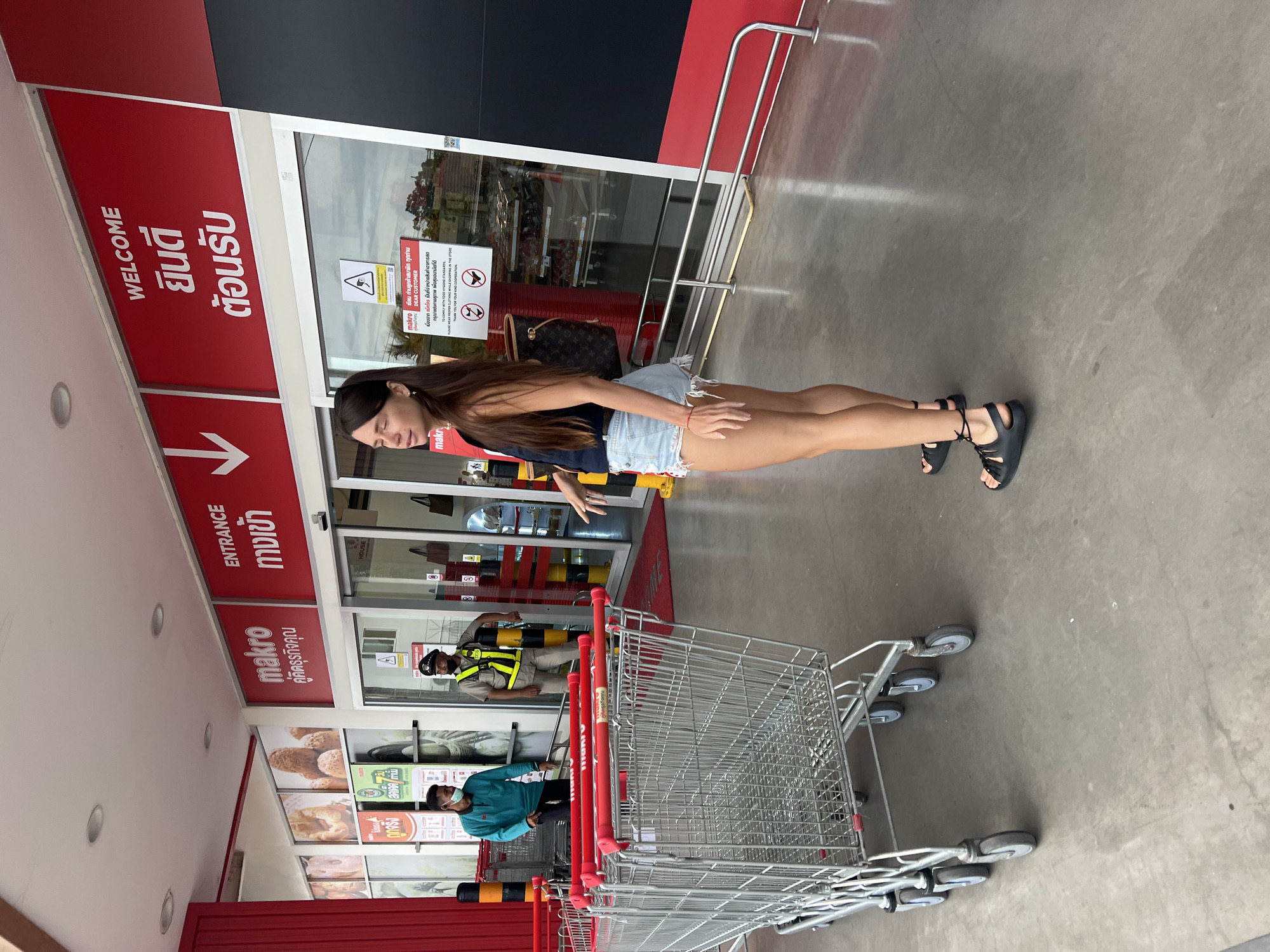 The image depicts the entrance area of a Makro store in Baan Hua Thanon, Thailand, during the daytime. A woman with long dark hair, wearing a dark sleeveless top, light denim shorts, and black sandals, stands in front of the automatic glass doors, looking downwards. Behind her, to the right of the frame, is a large stack of metal shopping carts.
Near the store entrance, a Louis Vuitton handbag is placed on the ground. Through the glass doors, a male staff member in a yellow reflective vest and cap stands by a stanchion, and another man in a blue shirt and face mask is visible further inside the store. The building's facade is predominantly red and black, with white signage.
Several signs are visible. A large vertical red sign to the left of the entrance reads "WELCOME ยินดี ต้อนรับ" (Welcome) and "ENTRANCE ทางเข้า" (Entrance) with an arrow pointing right. Above the entrance, another sign displays "makro คู่คิดธุรกิจคุณ" (Makro, Your Business Partner). On the glass door, a warning sign with the "makro" logo addresses "DEAR CUSTOMER" and advises them "TO COMPLY WITH FOOD HYGIENE STANDARDS, PLEASE WEAR PROPER CLOTHING WHILE SHOPPING IN THE STORE. THANK YOU FOR YOUR KIND COOPERATION." Inside, through the glass, images of bakery products are visible, with text reading "สดเดด เท่านั้น" (Fresh only) and "HOUSE". The ground is a light gray concrete.