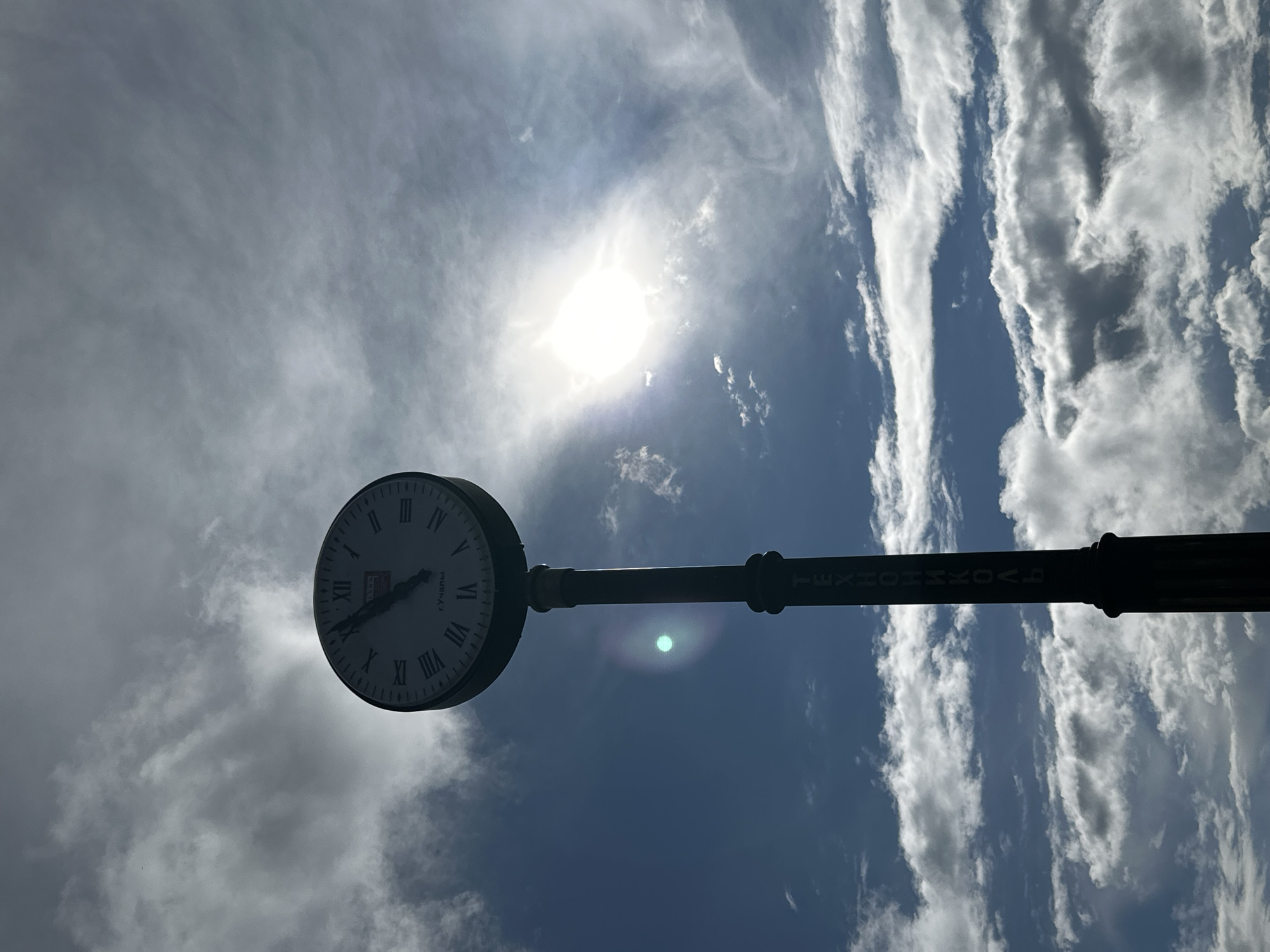 An outdoor shot features a street clock against a bright, partly cloudy sky. The image is oriented vertically, with the clock positioned on the left side, slightly below the center. The sun is prominently visible in the upper right quadrant, emitting a strong, bright light and creating a lens flare near the clock's pole. The sky is a mix of bright blue and scattered white and gray clouds, which appear thin and wispy around the sun and more defined towards the top and bottom edges of the frame.
The street clock is round with Roman numerals (I to XII) marking the hours. Its hour hand points towards VI, and the minute hand points towards X, indicating approximately 5:50. The clock face appears to have some Cyrillic text, including "ГУМ" (GUM) near the top and "ЦУМ" (TSUM) and "ТЕХНО" (TEKHNO) towards the right side. The clock is mounted on a dark, thick pole that extends horizontally across the middle of the frame towards the right. Along this pole, written vertically (when viewed in the image's original orientation), is the Cyrillic text "ТЕХНОСАД" (TEKHNOSAD). The entire clock and pole are silhouetted against the bright sky, emphasizing the sun's intensity. The scene captures a bright daytime, possibly late morning or early afternoon.
