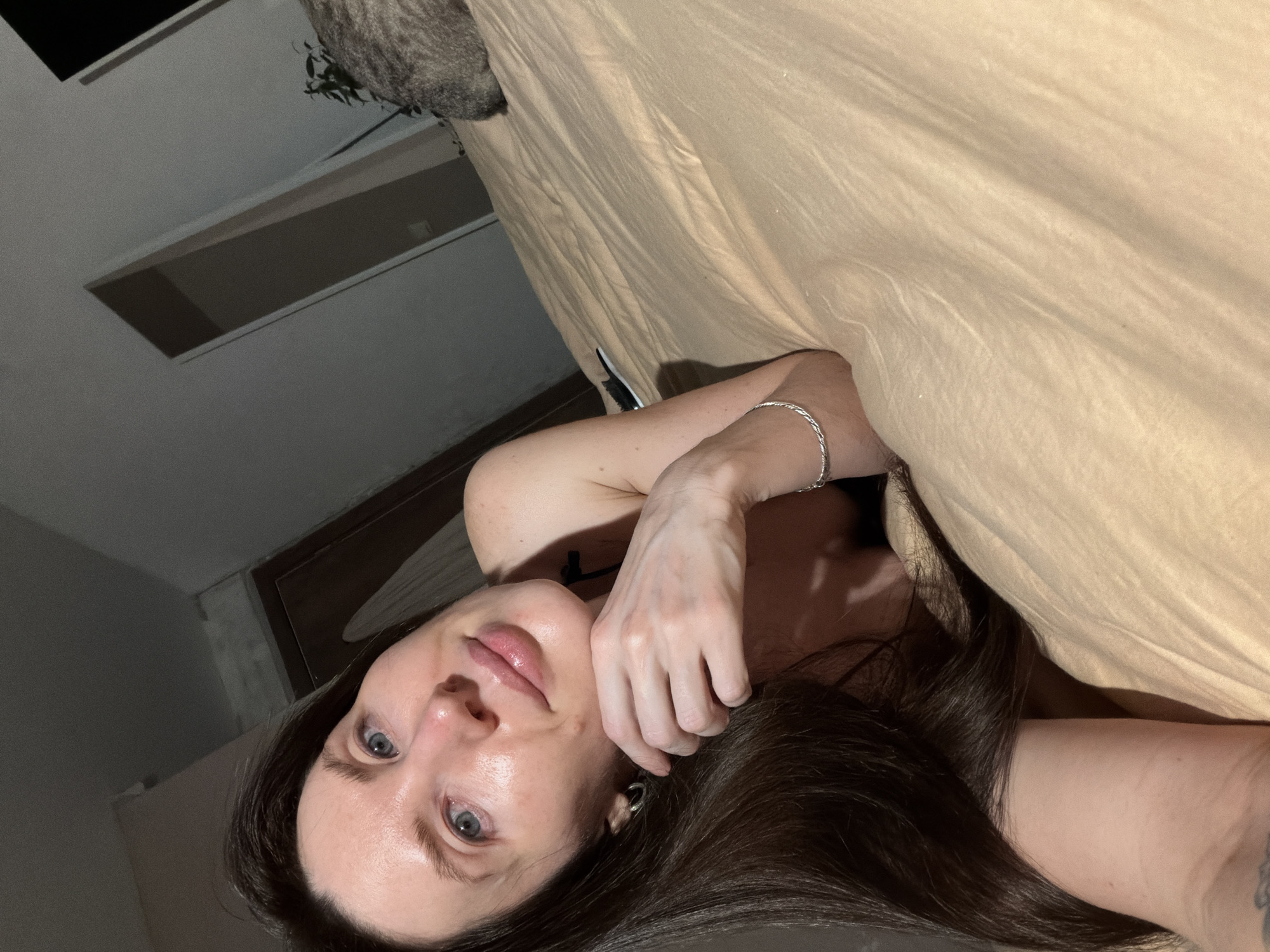 A woman with long, dark brown hair is lying in bed under a beige blanket. She has fair skin and blue eyes, and is wearing a silver bracelet on her left wrist. The background includes a window and a door or doorway, suggesting an indoor setting. The location is Perm, Russia.