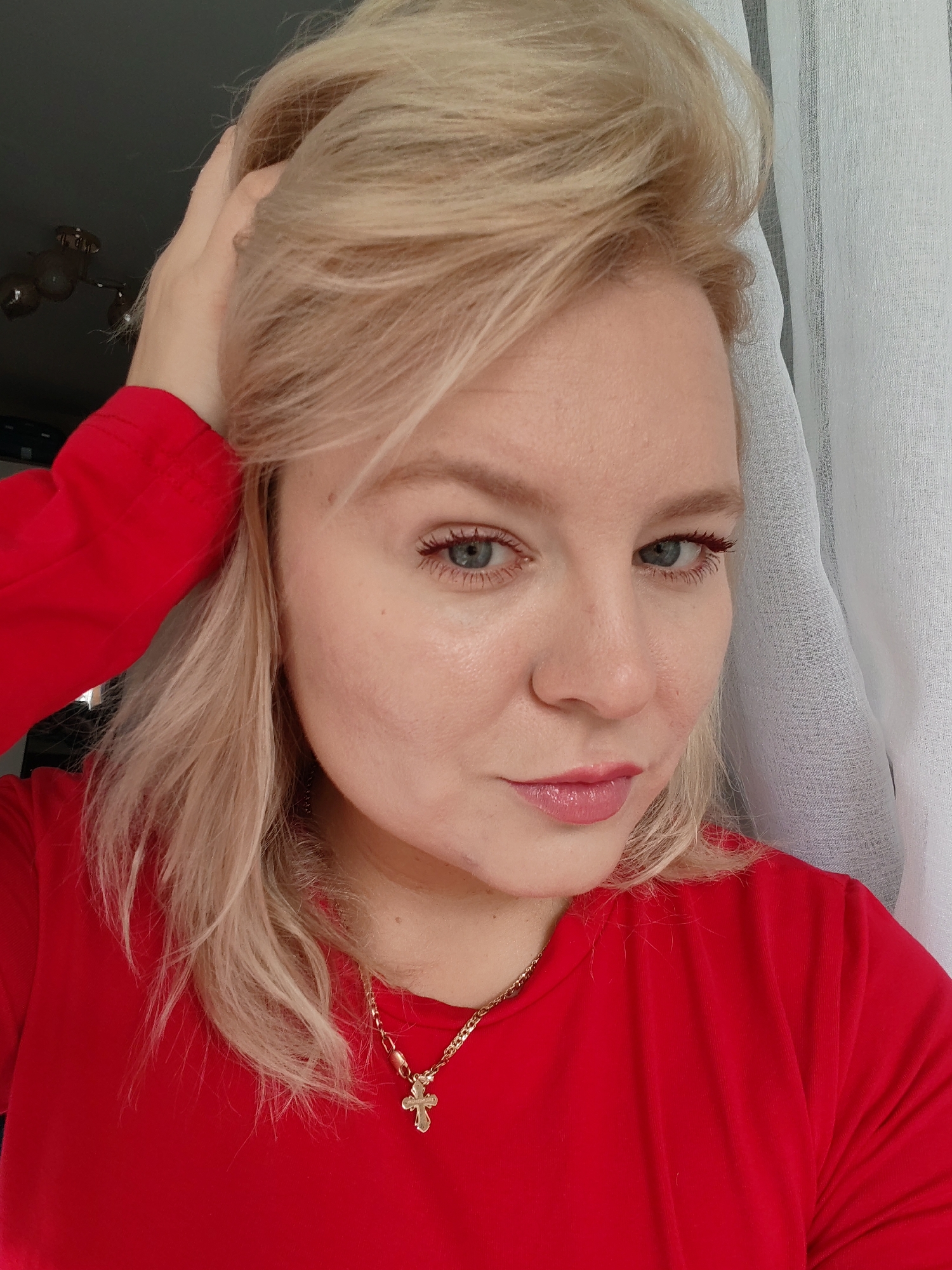 The image is a close-up, waist-up shot of a fair-skinned woman with blonde hair, wearing a red long-sleeved shirt and a gold chain with a cross pendant. She is looking towards the camera, with her left hand touching her hair. The background features a white curtain with vertical stripes and a dark area that appears to be an interior space, possibly a room in Izhevsk, Russia. The lighting suggests it is daytime.