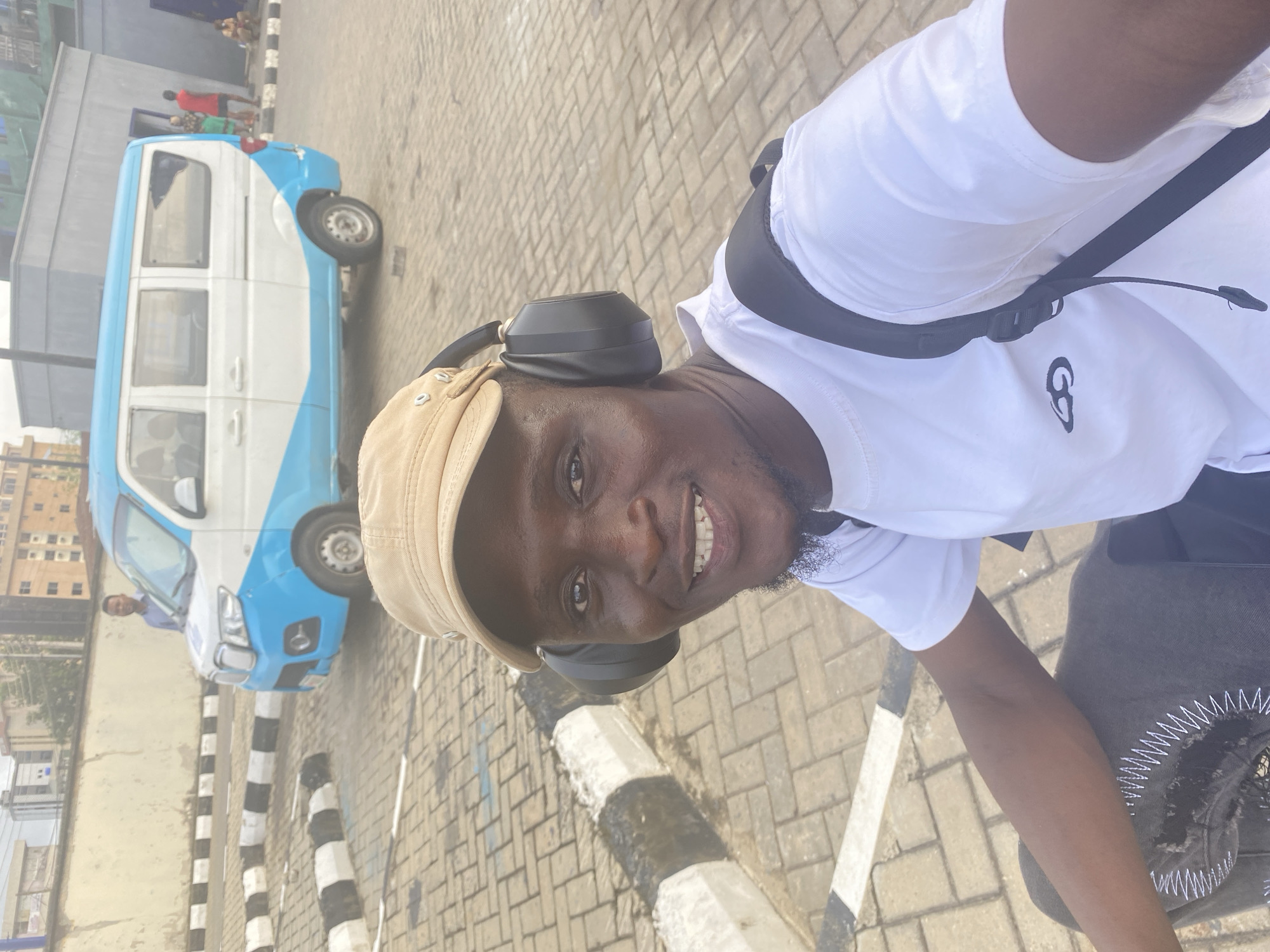 The main subject of the image is a smiling Black man, appearing to take a selfie or be photographed outdoors in an urban setting. He looks directly at the camera with a cheerful expression, showing his teeth. He wears a light brown or beige cap, black over-ear headphones, and a white short-sleeved t-shirt with a black, stylized 'G' or abstract circular logo on the left chest. A black strap from a sling bag is visible across his chest.
The scene depicts a paved street or roadside in Ikeja, Nigeria, under bright daylight, indicating clear, sunny weather. The ground is covered with light-colored interlocking brick pavers. In the mid-ground, a blue and white passenger minibus, characteristic of public transportation vehicles in the region, is parked or moving. An adult male is partially visible near the front of the minibus. Black and white striped concrete curbs or barriers delineate the road edge. Further in the background, various multi-story buildings suggest a bustling urban landscape. The man's smile and the bright lighting contribute to a positive and vibrant atmosphere.