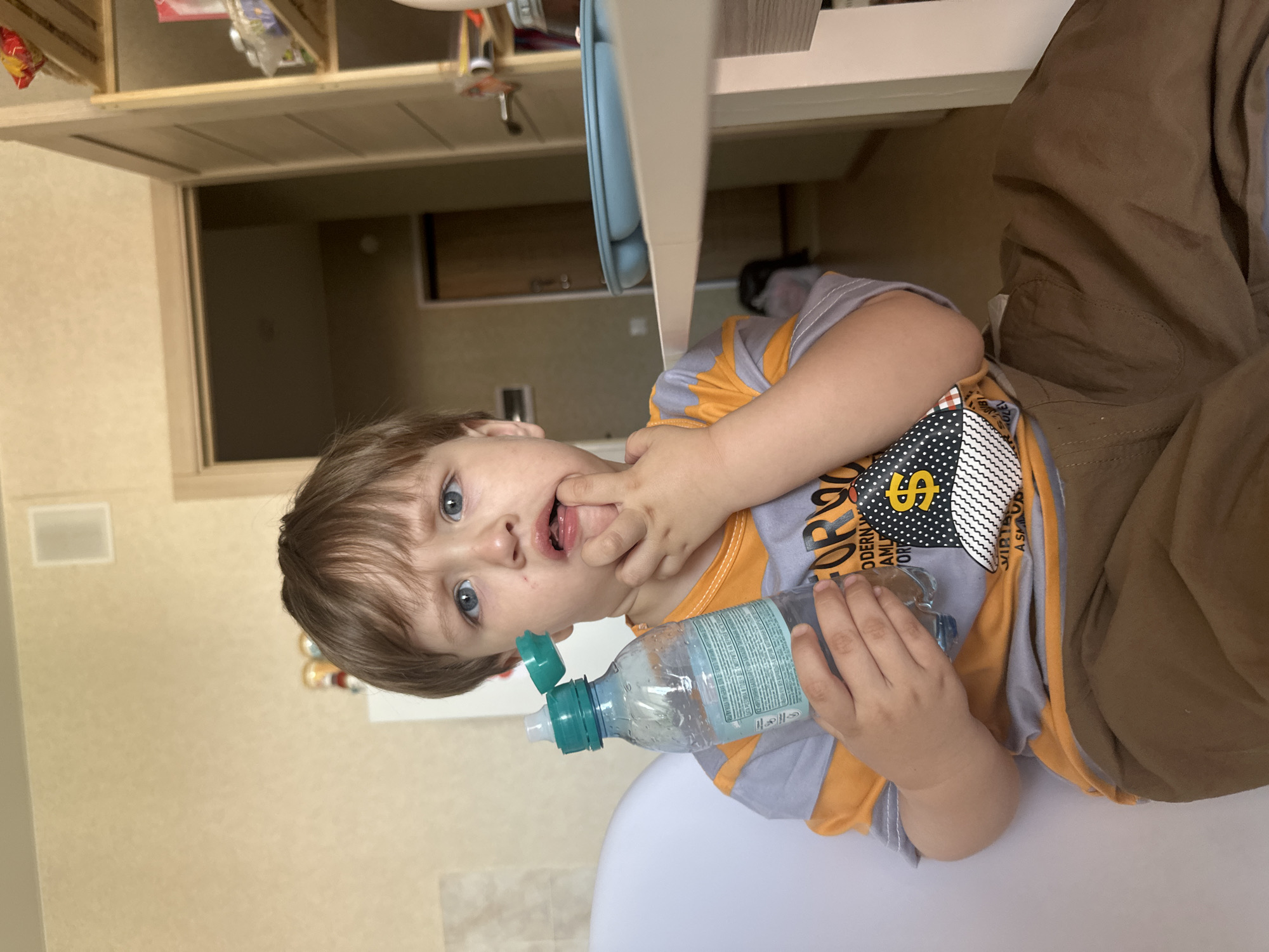 A young child, appearing to be of Caucasian descent with brown hair and blue eyes, is seated and holding a plastic water bottle. The child's left hand is near their mouth, with a finger touching their lips. They are wearing a t-shirt with orange and grey stripes and a graphic design featuring a dollar sign on the front, along with brown pants. The background suggests an indoor domestic setting, possibly a kitchen or dining area, with a doorway visible in the distance and shelves filled with items. The overall lighting indicates daytime. The image does not contain any information that would allow for a precise location within Ussurijsk, Russia.