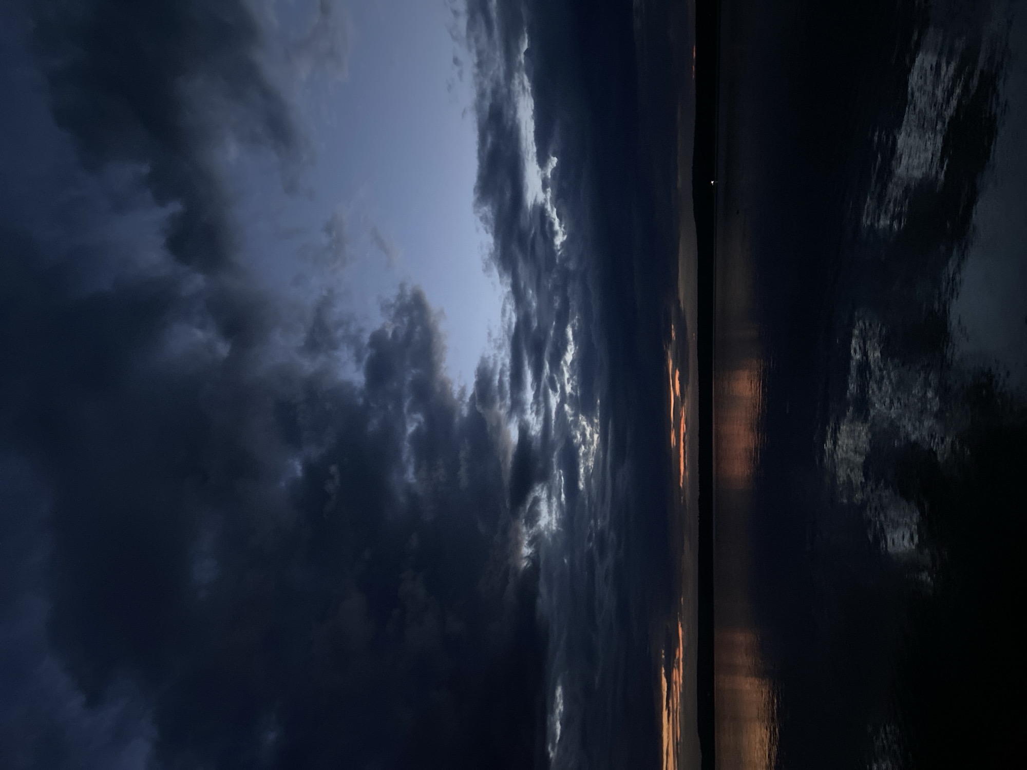 The image depicts a serene body of water, likely a lake or river, at dusk. The sky is filled with dramatic, dark clouds, with streaks of orange and yellow light peeking through near the horizon, suggesting the sun is setting. The water's surface is calm and reflective, mirroring the colors of the sky and the dark silhouettes of distant landforms. A faint light source, possibly a distant building or vessel, is visible on the far shore. The overall atmosphere is one of quiet contemplation and natural beauty. The location is identified as the city of Votkinsk, Russia. There are no people or discernible objects in the foreground or middle ground.