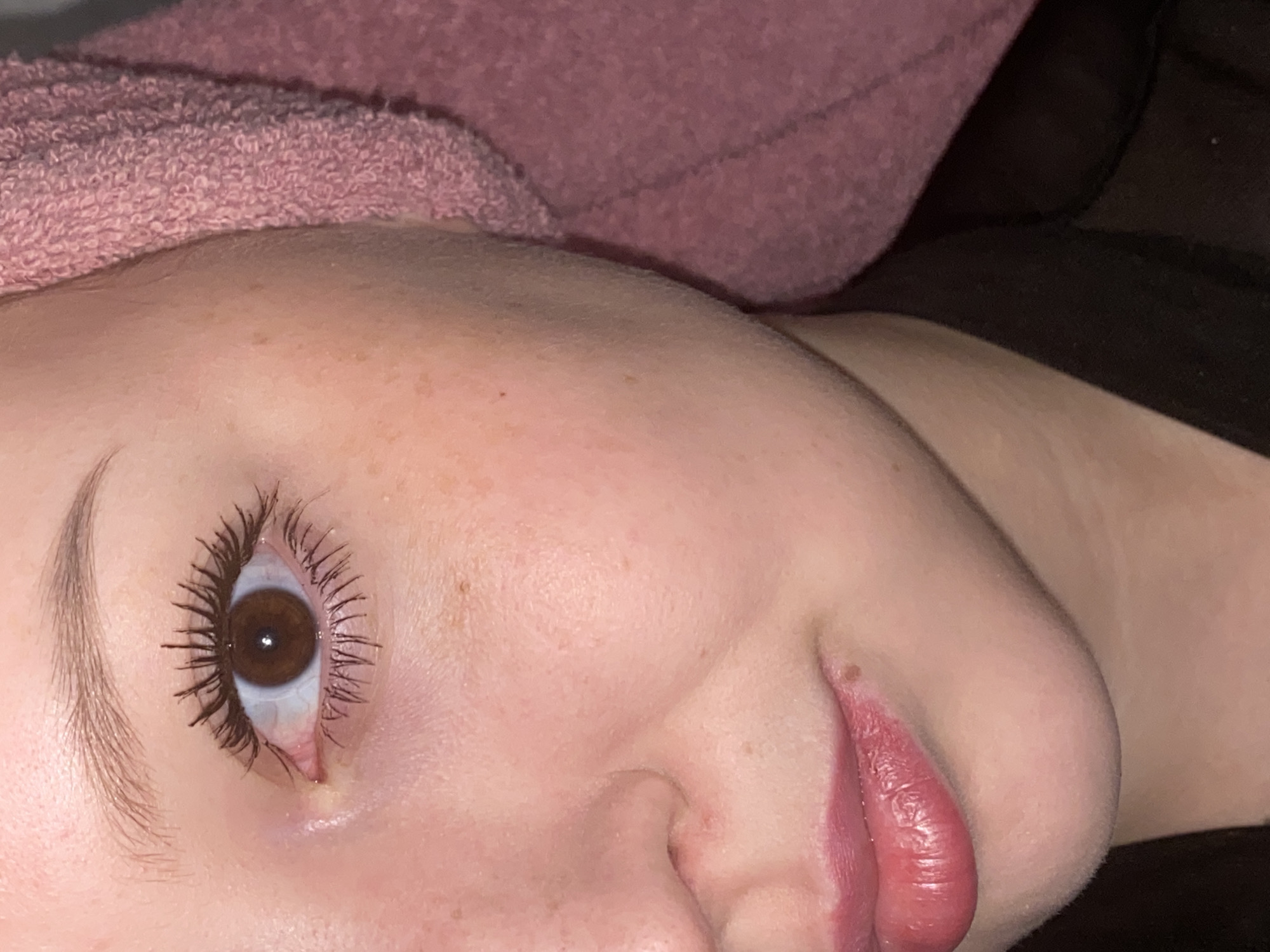 A close-up, angled shot of a person's face. The individual has fair skin, brown eyes with mascara applied, and light eyebrows. A pink towel rests on their head. Their lips are visible, appearing moisturized. Freckles are scattered across the skin. The background consists of a dark, indistinct surface. No information is available from the provided image to suggest any location in Chaikovsky, Russia, or anywhere else.