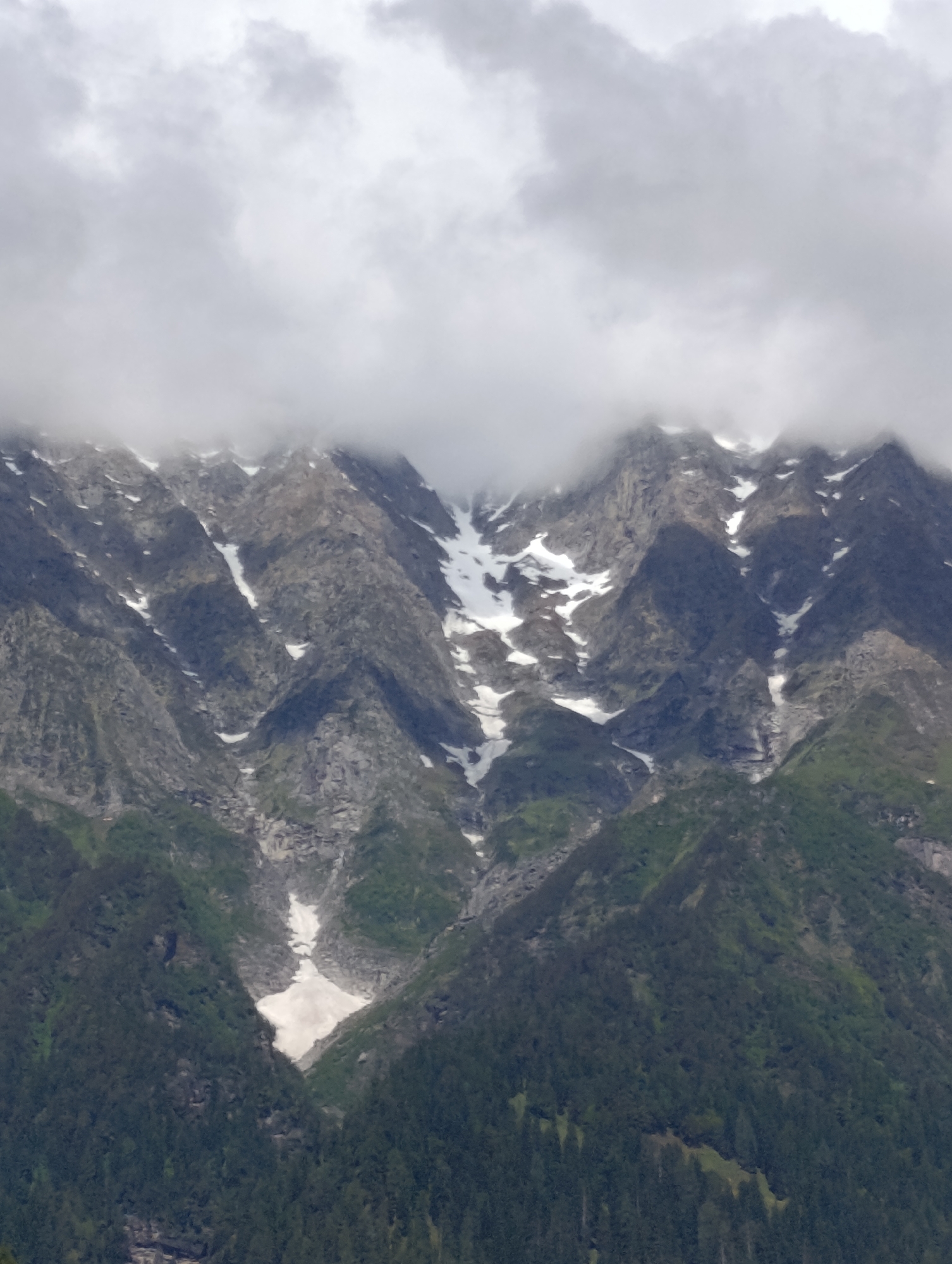 The image is a vertical, medium shot of a mountainous landscape with patches of snow and dense evergreen trees. The upper portion of the mountains is shrouded in thick, grey clouds, obscuring the peaks and creating a dramatic, atmospheric effect. The rocky slopes are a mix of dark grey and lighter, textured stone, with diagonal streaks of white snow running down their faces. The lower slopes transition into a verdant green, covered in a dense carpet of coniferous trees. A prominent snowfield is visible in a valley or crevice on the lower left side of the mountain, contrasting sharply with the surrounding dark foliage. The lighting is diffused and overcast, typical of cloudy mountain weather. The scene depicts a remote, natural environment within the city of Kullu, India. There are no people or animals visible in the image.