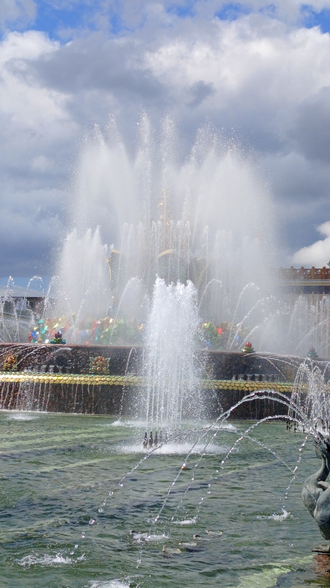 The image depicts a large, multi-tiered fountain in Moscow, Russia. The fountain is active, with numerous streams of water spraying upwards and outwards, creating a misty effect. The water is illuminated by sunlight, and droplets are visible in the air. The fountain basin is elaborately decorated with what appear to be mosaic or tile work featuring floral patterns and gold trim. In the foreground, the water is a deep green color, with smaller arcs of water falling into it. A statue of a bird or swan is partially visible in the lower right corner. In the background, a building with ornate architecture is visible, along with a cloudy blue sky. The overall scene suggests a sunny but potentially windy day in a public park or square.