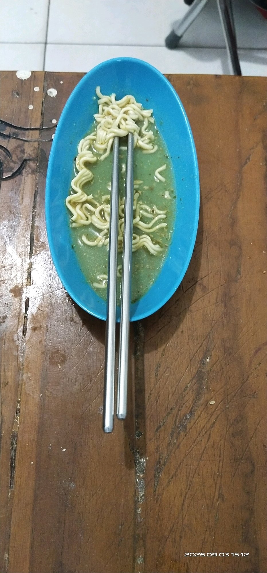 A close-up, top-down shot shows a blue oblong bowl filled with instant noodles in a green broth, resting on a dark wooden table. Two metallic chopsticks are placed vertically in the bowl, holding some of the noodles. The table has a rough texture with visible grain and some scuff marks. In the background, the edge of a tiled floor is visible on the left, and a tripod-like object can be seen blurred in the upper right corner. The timestamp "2026.09.03 15:12" is overlaid on the bottom right of the image. The scene appears to be indoors, likely a home setting, possibly in Tamiang Layang, Indonesia, given the context provided.